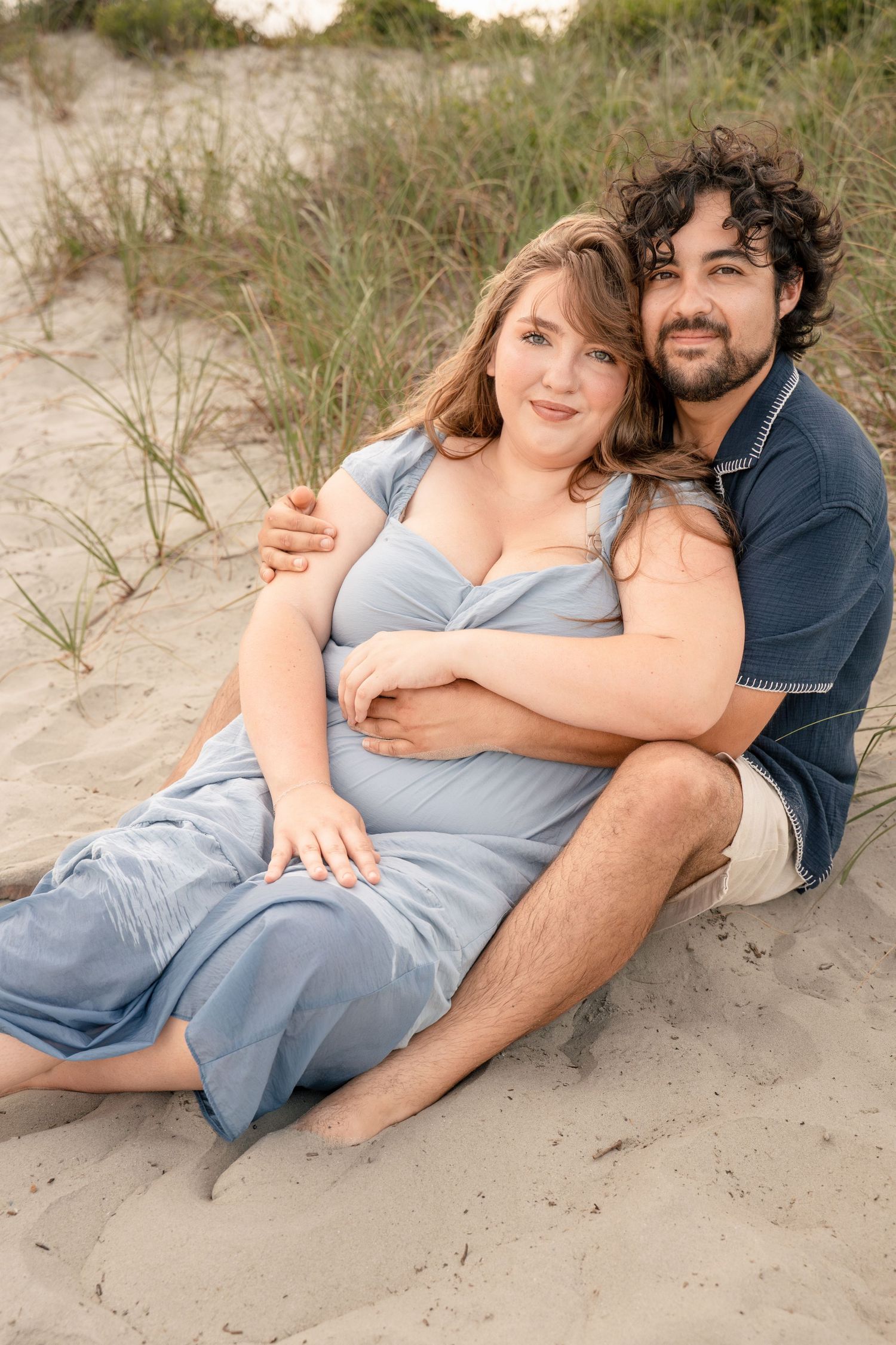 A couple sits together on sandy beach dunes, smiling warmly while embracing near coastal grasses.