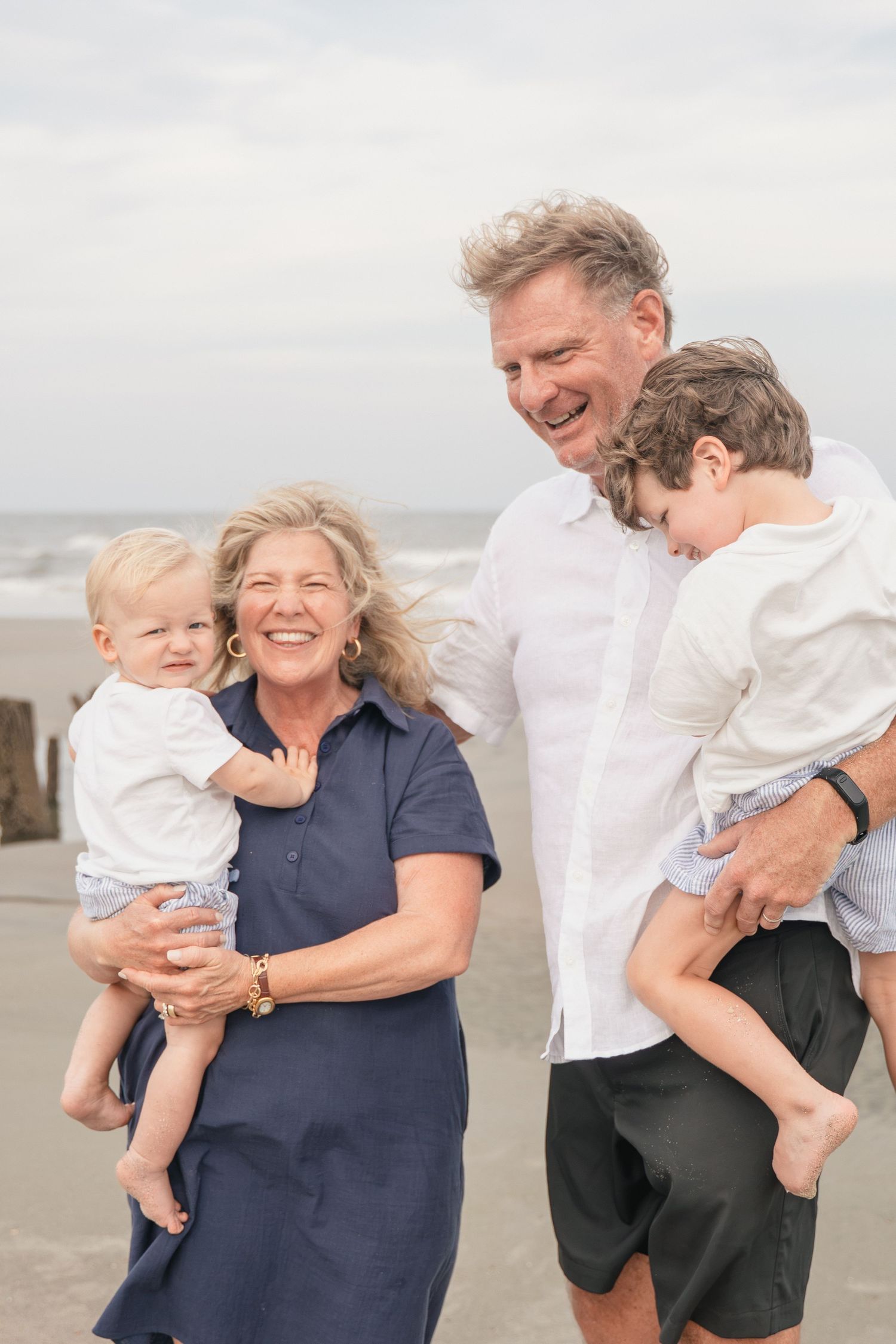 Grandparents laughing and holding two young grandchildren on a beach with ocean waves in the background.