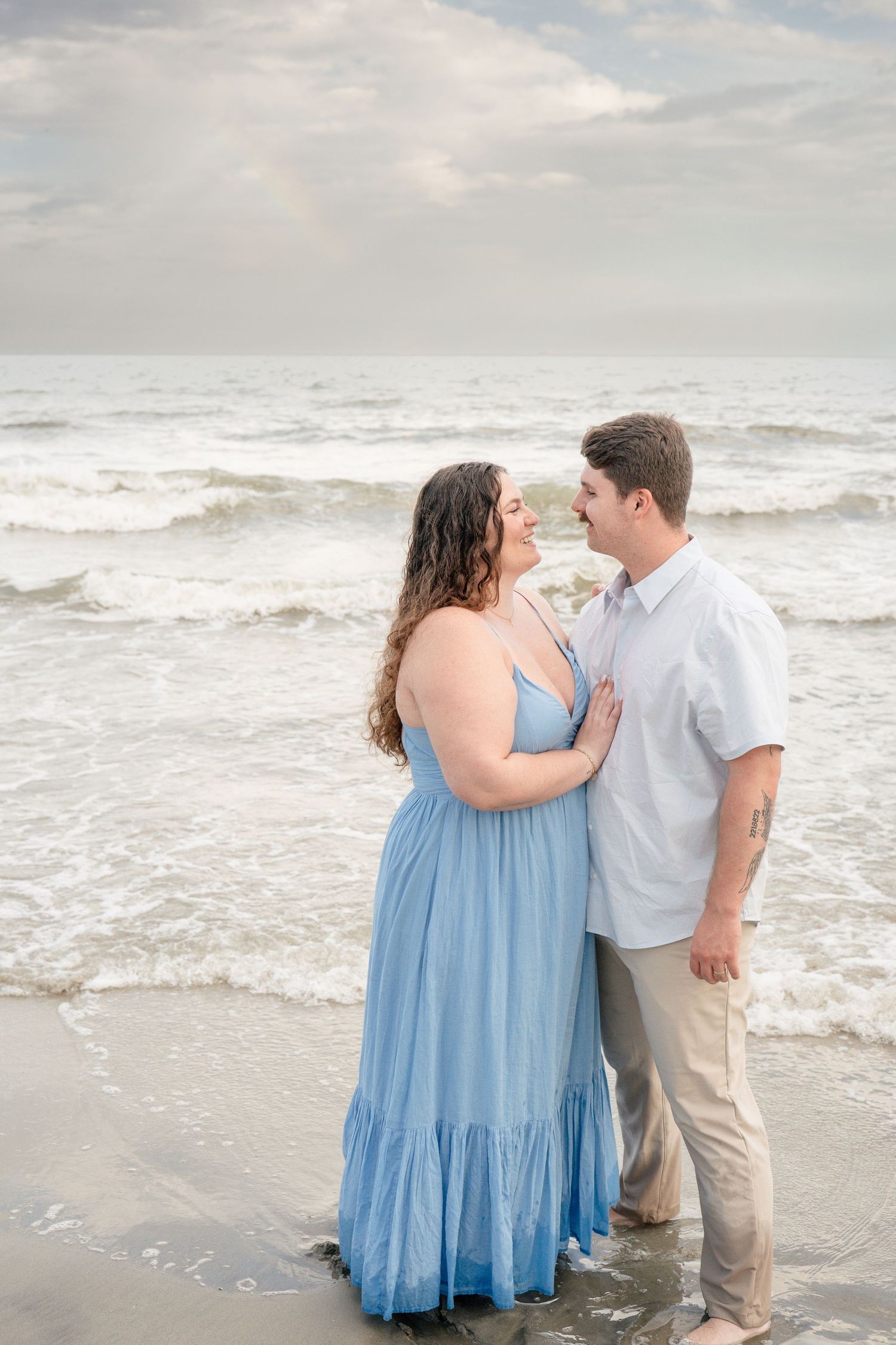 Couple in blue dress and white shirt embrace on a beach with ocean waves behind them.