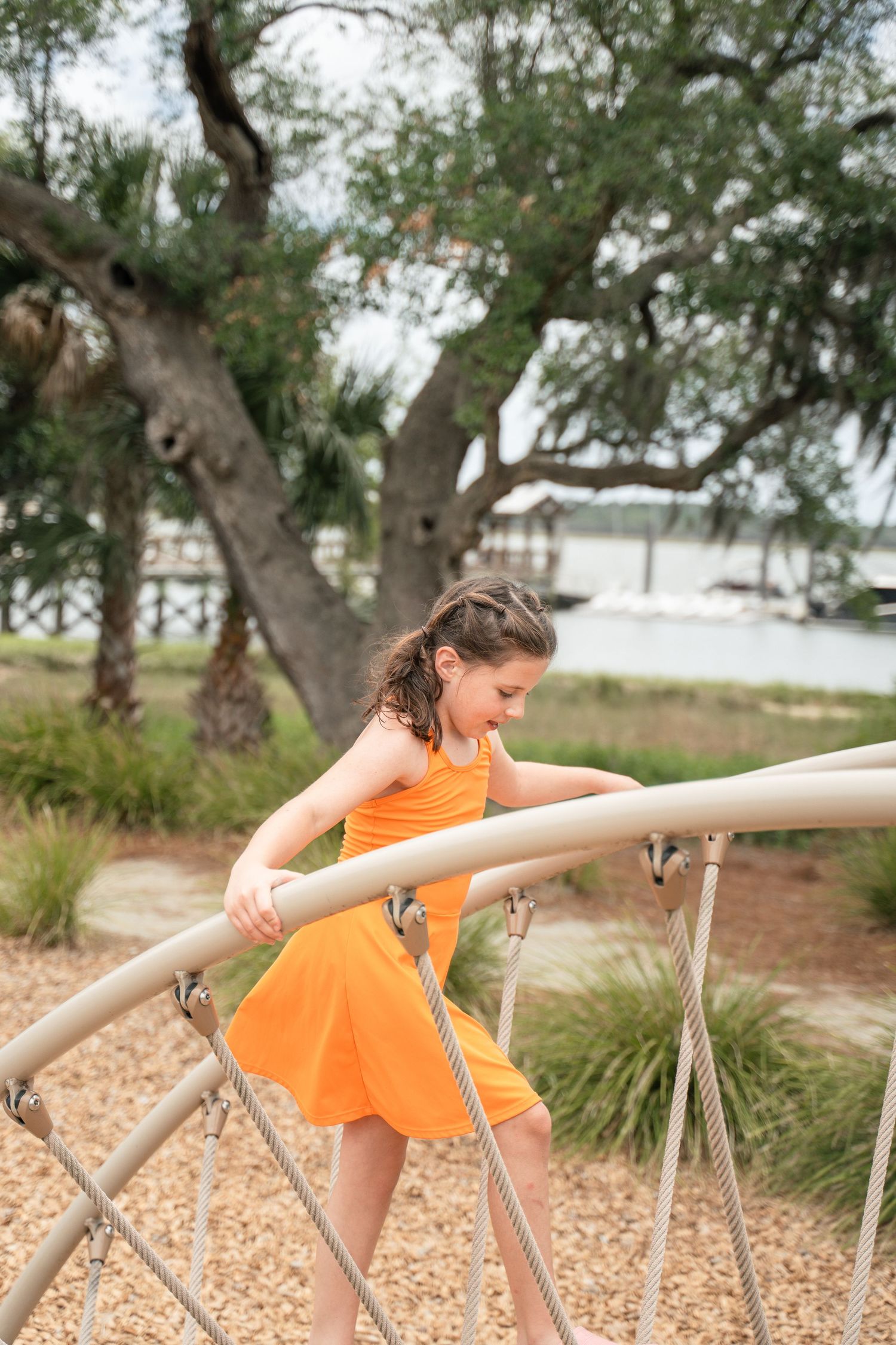 A young girl in an orange dress climbs a rope structure at an outdoor playground near a waterfront park.