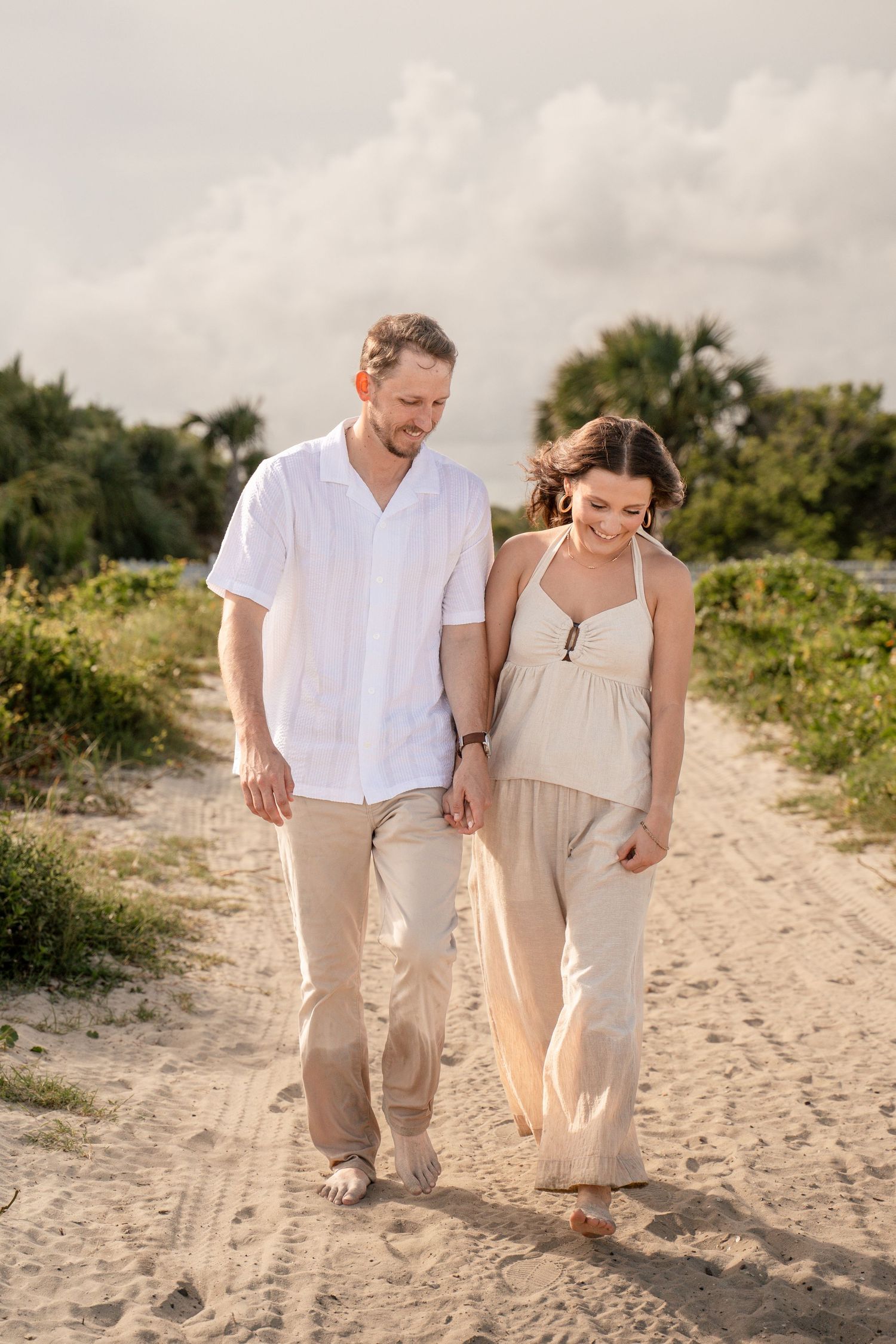 Couple walking hand-in-hand along a sandy beach path surrounded by tropical greenery at sunset.