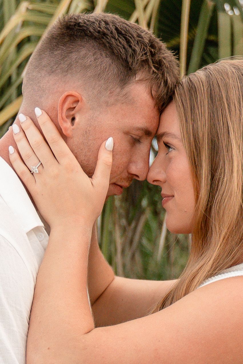 Couple sharing an intimate forehead touch, woman displaying engagement ring, with tropical palm leaves in background.