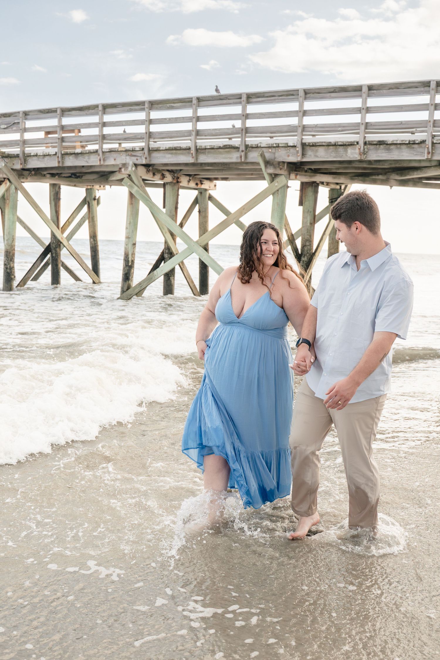Couple holding hands walking in ocean waves near a pier, woman in flowing blue maxi dress, maternity beach photo session.