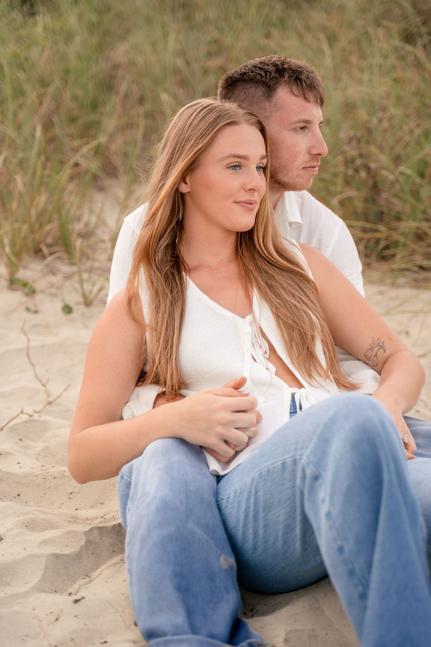 Young couple sitting together in beach sand dunes, woman leaning back against man in casual attire.