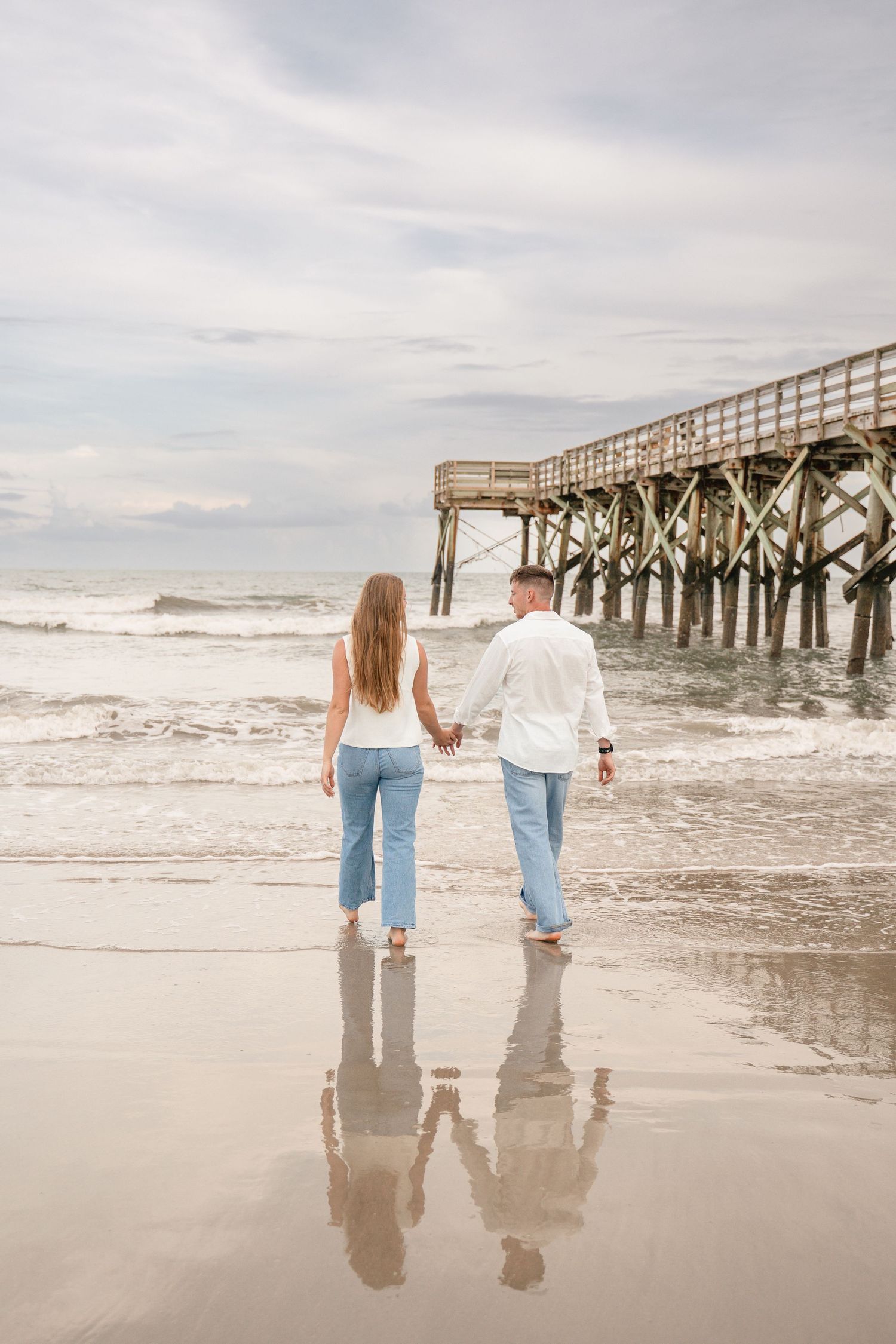 Couple holding hands walking barefoot on a reflective beach near a wooden pier with ocean waves.