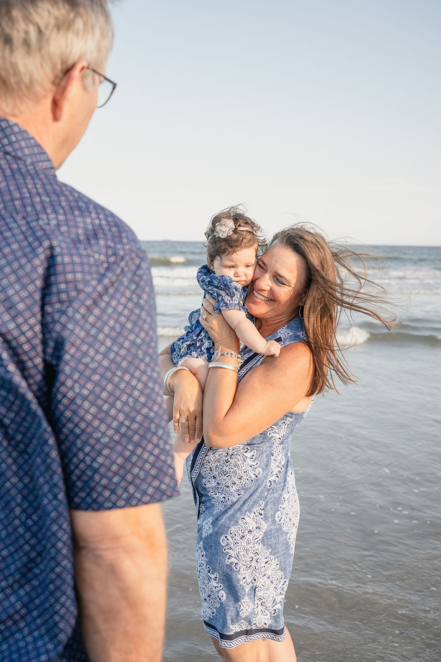 Grandparents smiling and holding a baby girl on a sunny beach with ocean waves in the background.