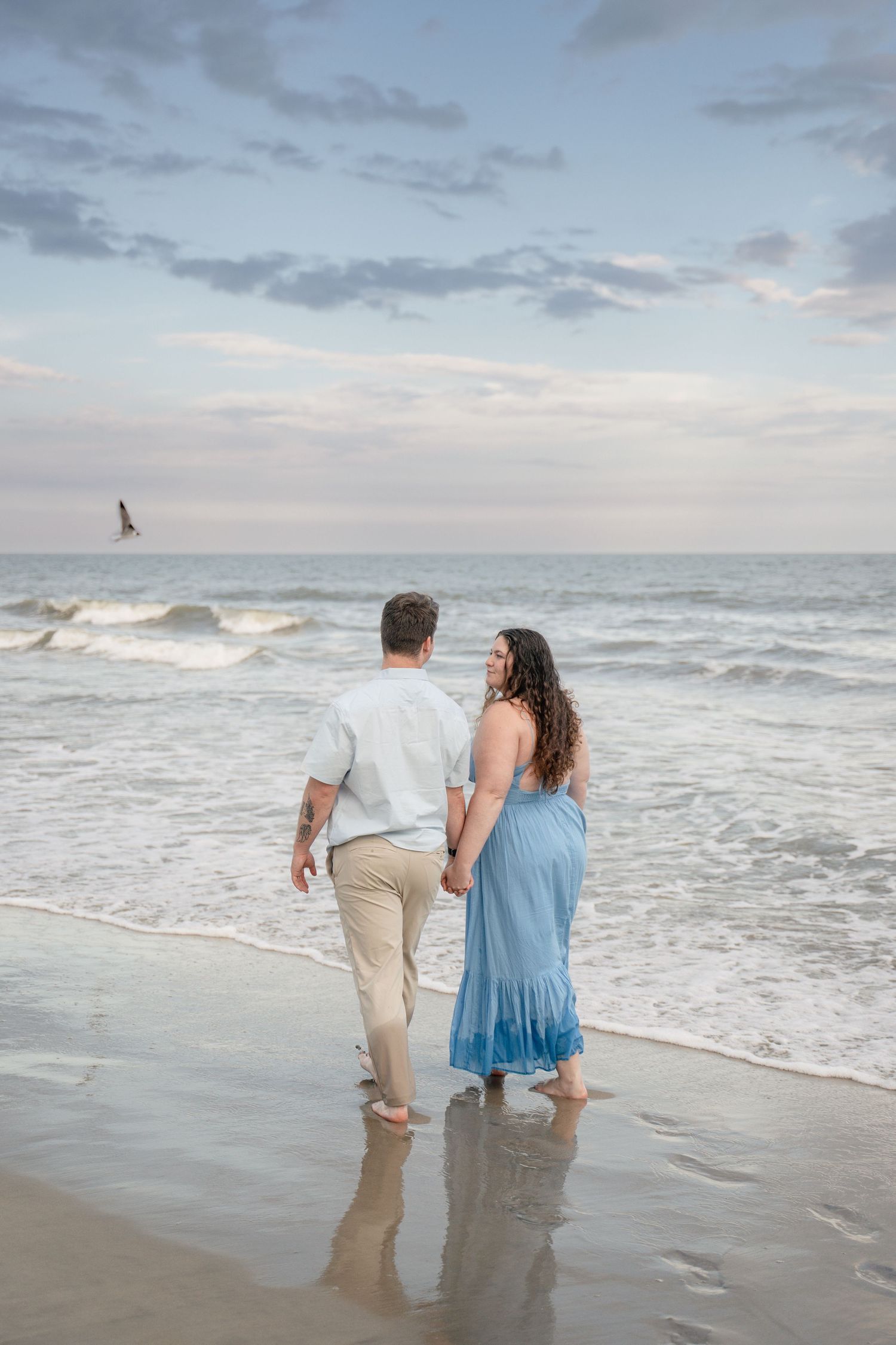 Couple holding hands walking barefoot along ocean shoreline, woman in blue maxi dress, man in white shirt and khaki pants.