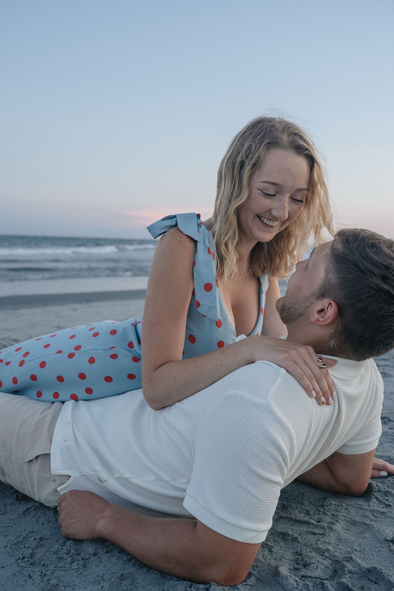 Couple sharing a romantic moment on the beach at dusk, woman in polka dot dress smiling at man lying on sand.