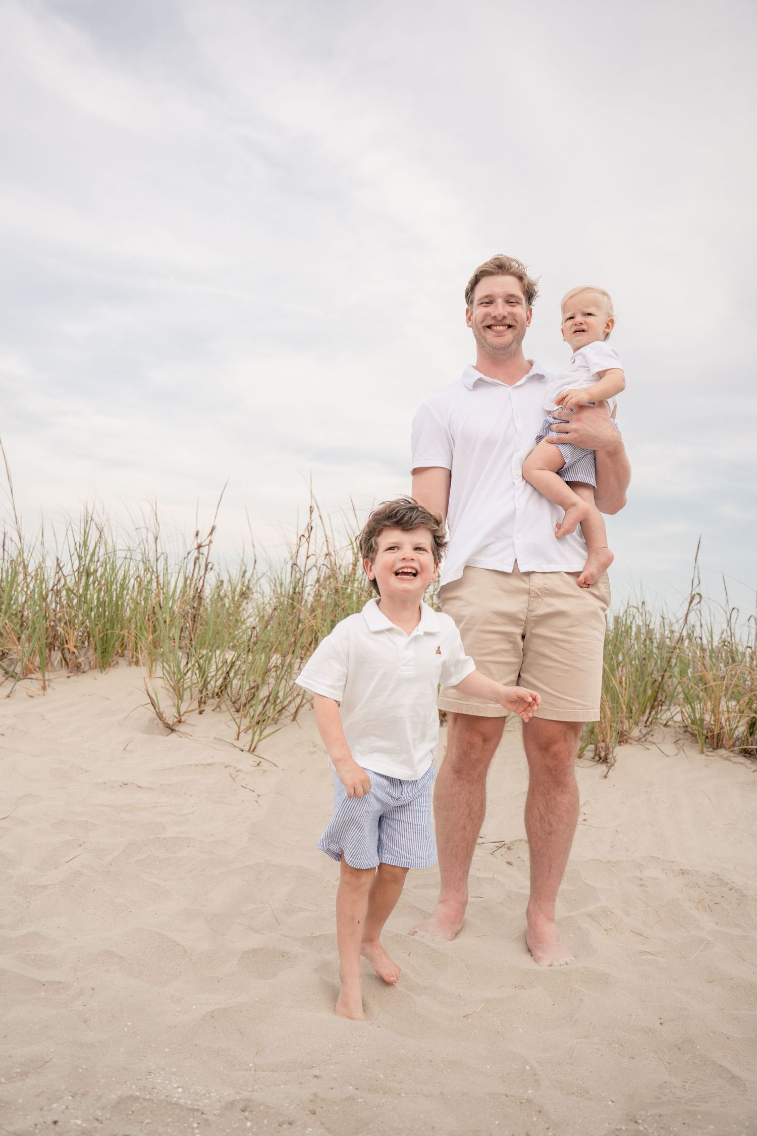 Father smiling at beach with two young sons, holding baby while older boy laughs on sandy dunes.