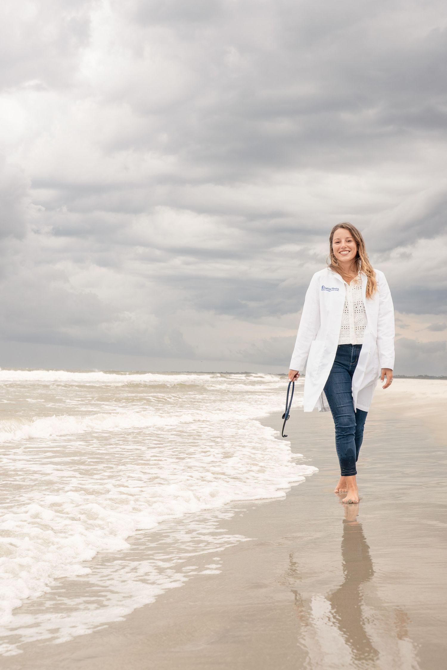 Woman in white coat and jeans walking barefoot on a sandy beach, holding shoes, smiling under cloudy skies.