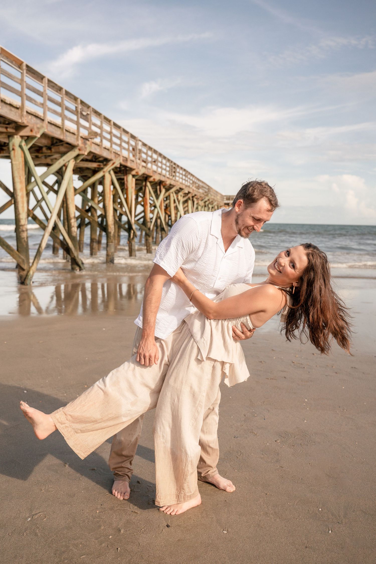Man dipping laughing woman in beige outfit on a reflective beach near a wooden pier.