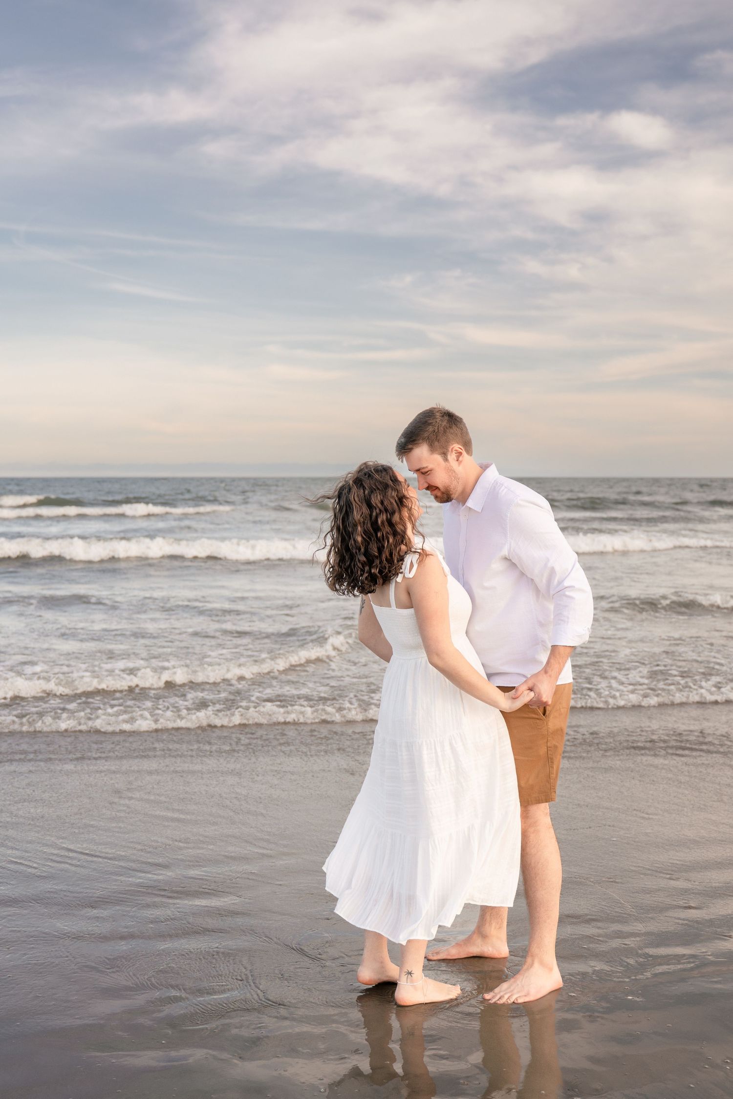 A couple stands barefoot on a wet beach shore, gazing at each other, woman in white dress, man in white shirt and tan shorts.