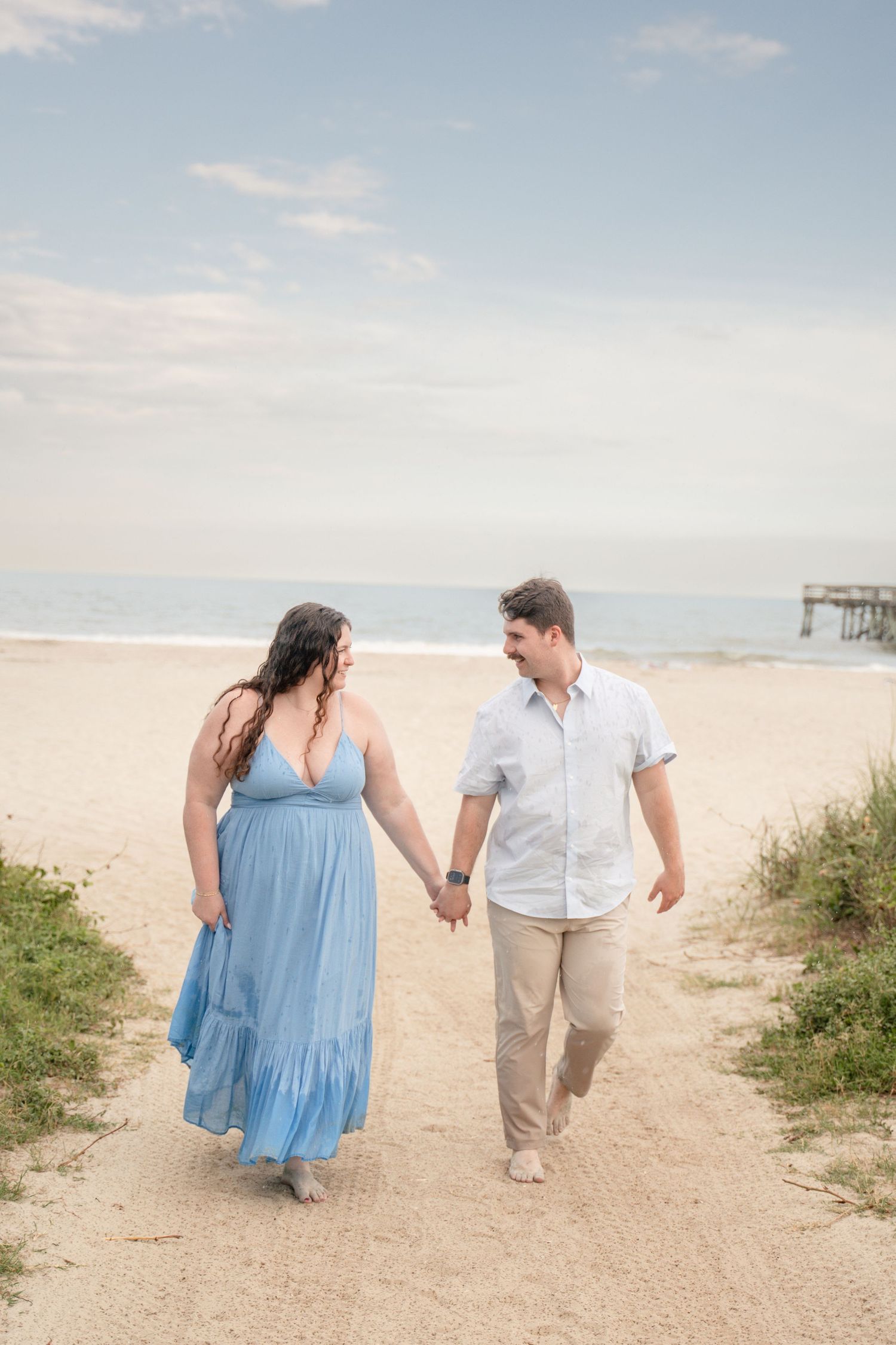 Couple holding hands walking on sandy beach path, woman in blue maxi dress, man in white shirt and khaki pants.