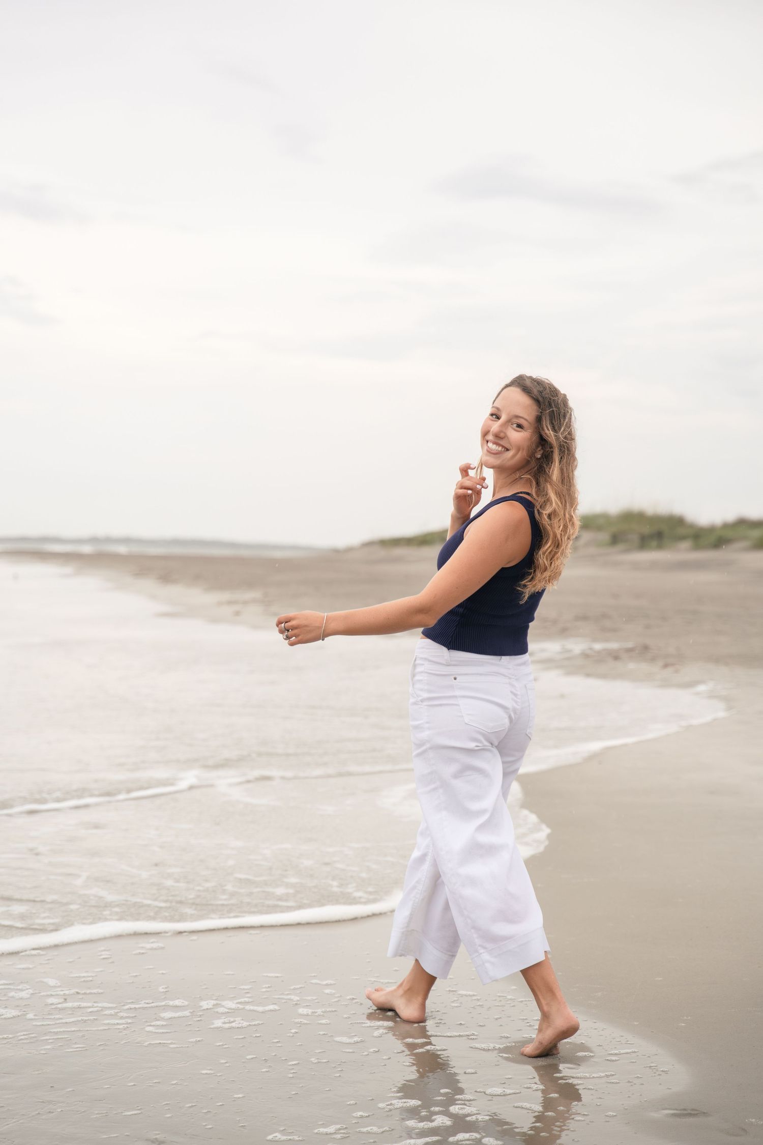 A smiling woman in white pants and navy top walks barefoot along a sandy beach shoreline.