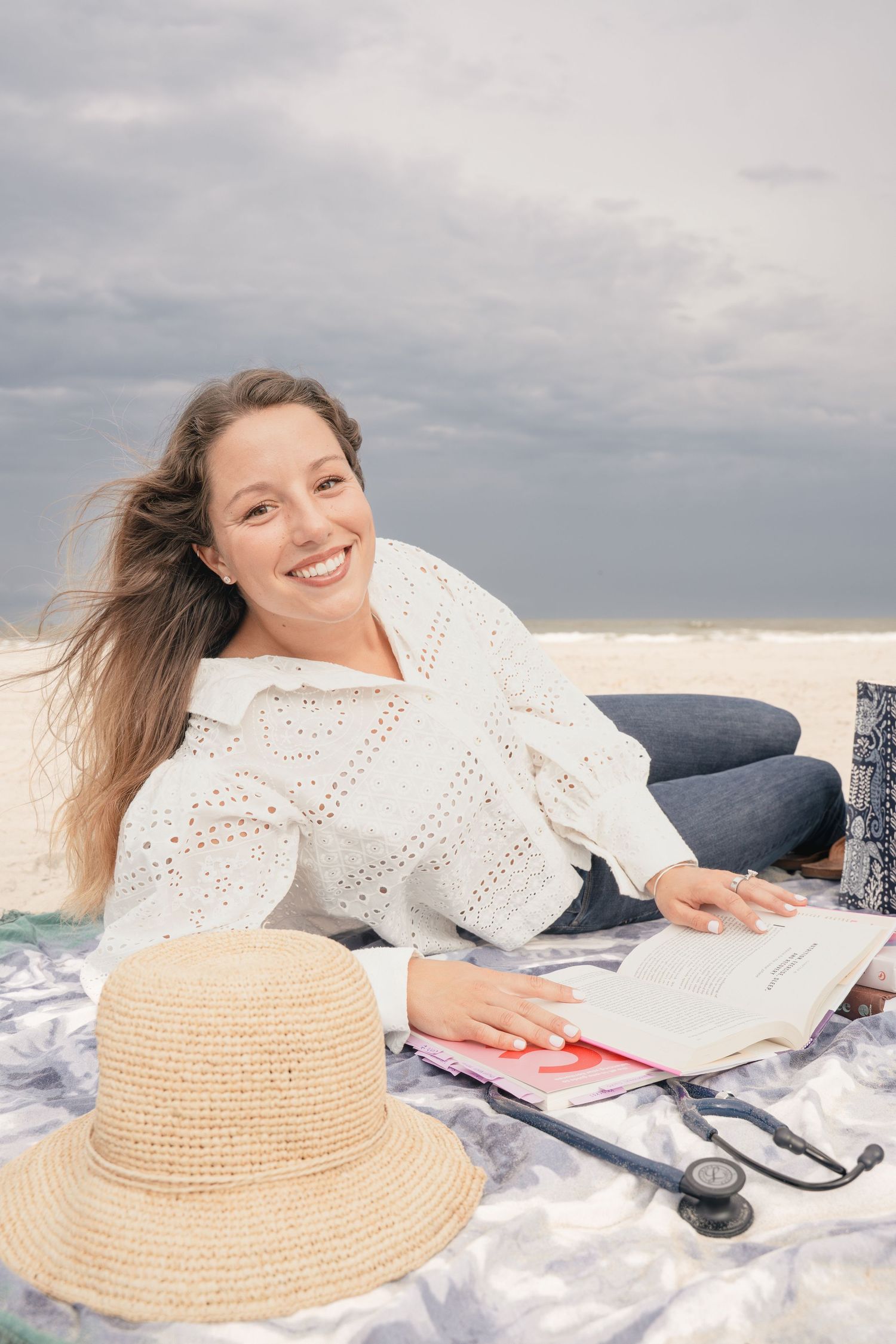 Smiling woman in white sweater lying on beach blanket with straw hat, stethoscope, and open book under cloudy sky.