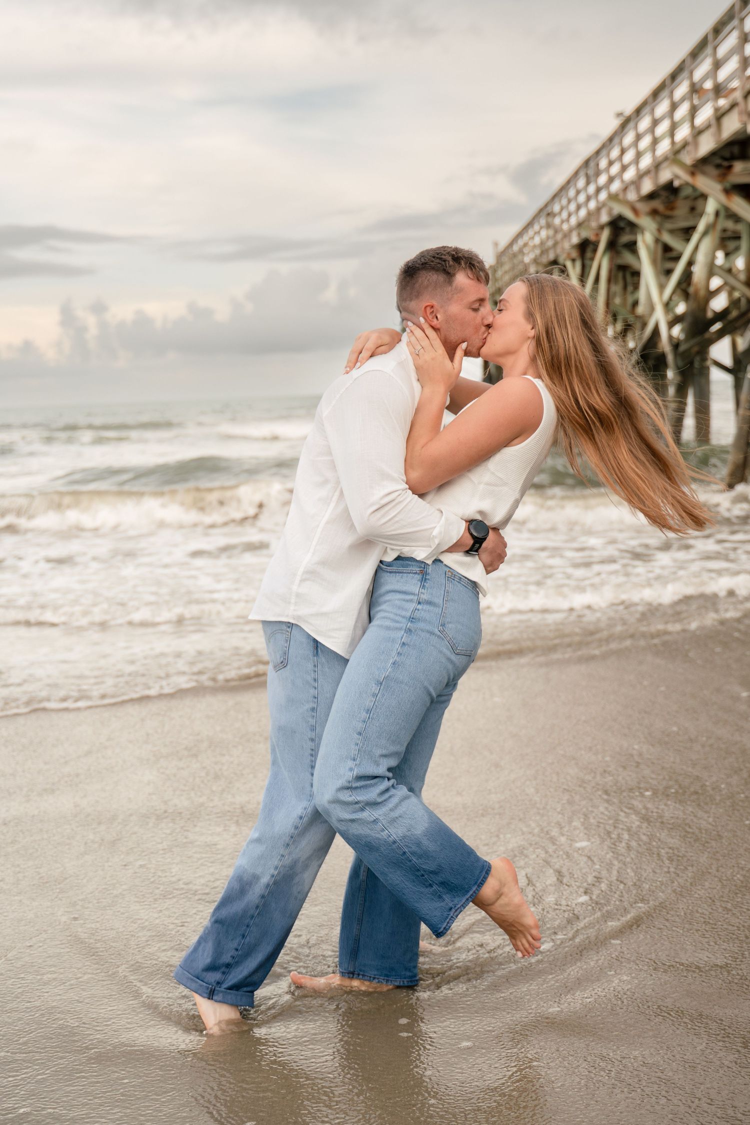 Couple sharing a passionate kiss in the ocean waves on the beach beside a wooden pier.