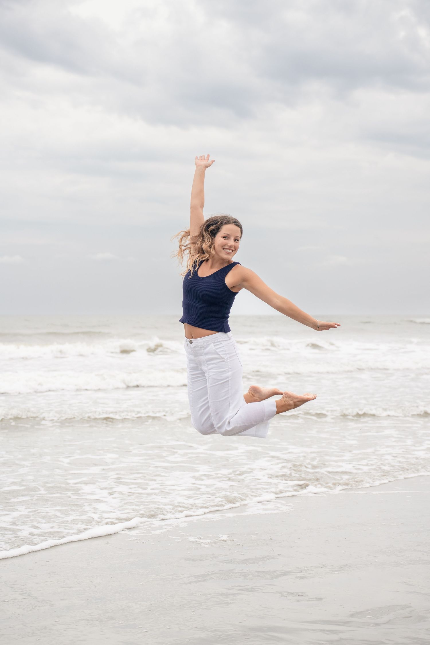 A joyful young woman in white pants and navy top jumping on a beach with ocean waves in the background.