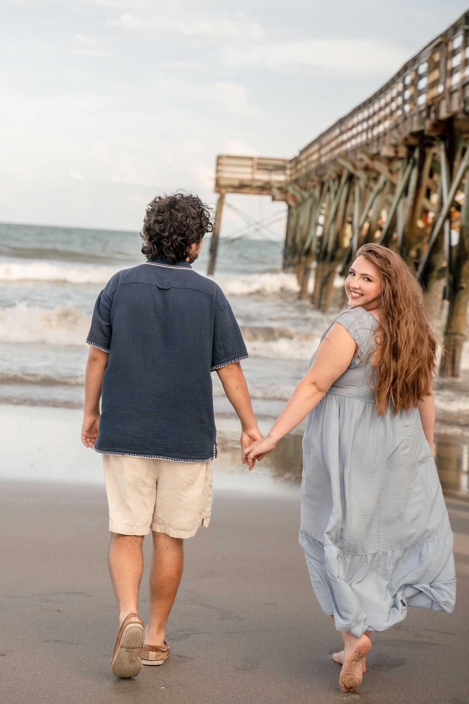 Woman in blue dress looking back smiling while holding hands with man walking toward ocean pier.