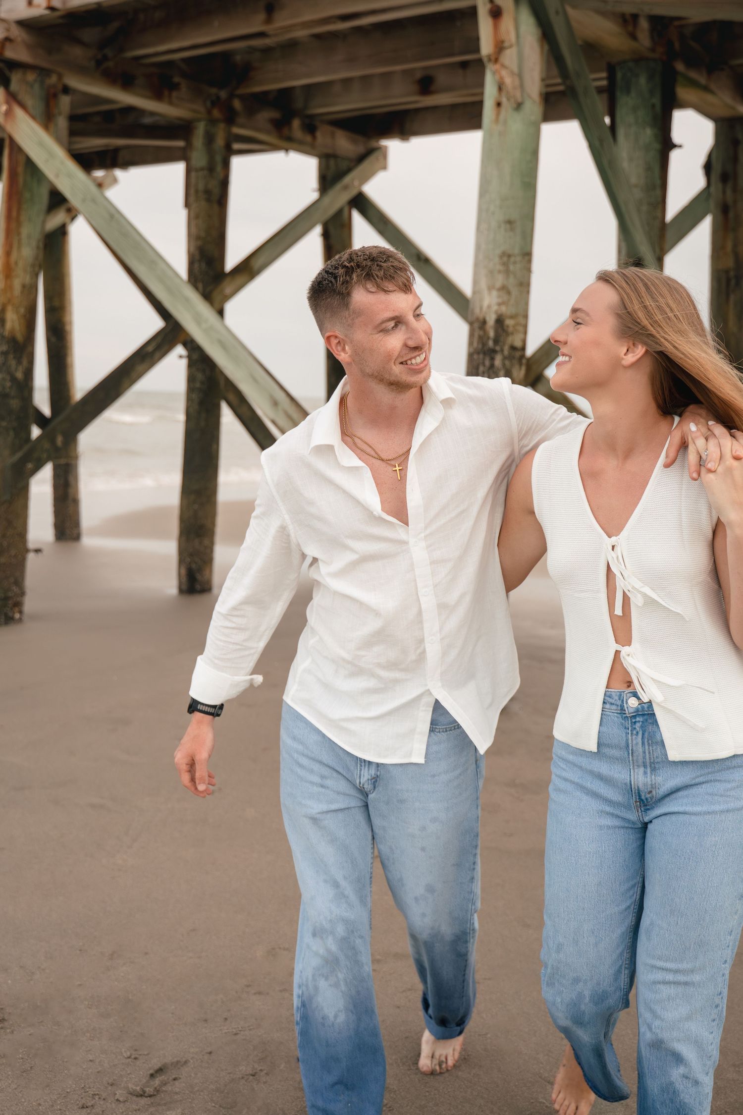 A couple in matching white shirts and jeans walk and laugh together on a sandy beach near a wooden pier.
