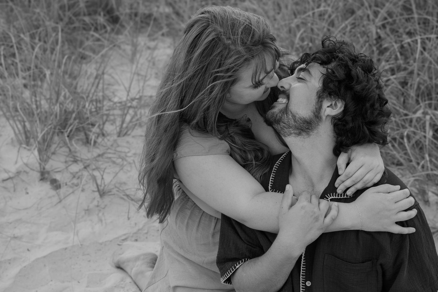Black and white photo of a romantic couple embracing and kissing on a sandy beach with dune grass.