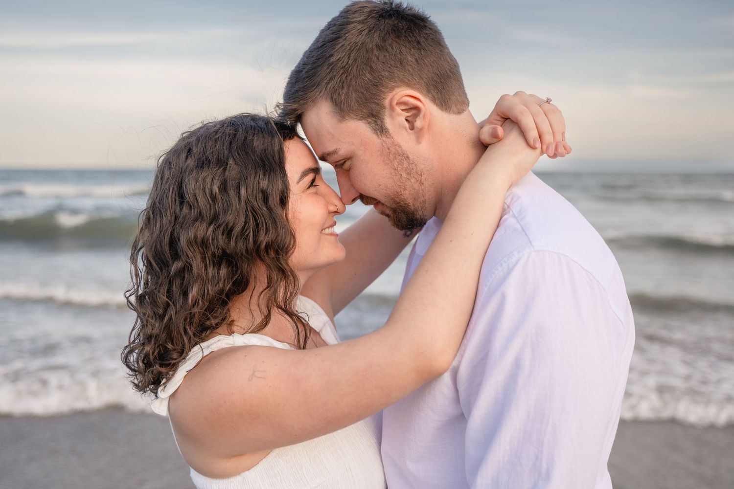 Romantic couple embracing on a beach at sunset, touching foreheads and smiling near ocean waves.