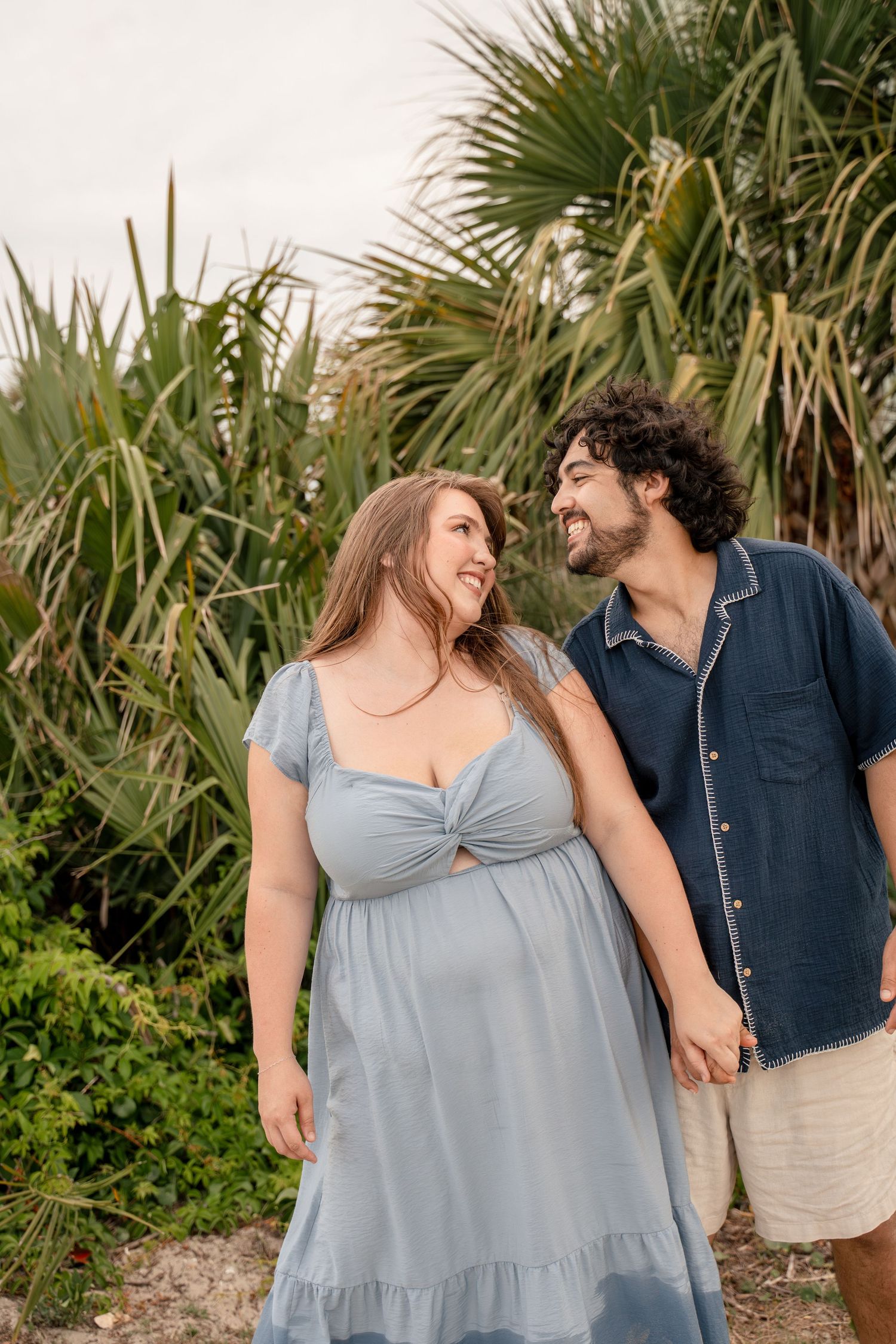 A smiling couple holds hands and walks together in front of lush tropical palm trees on a beach.