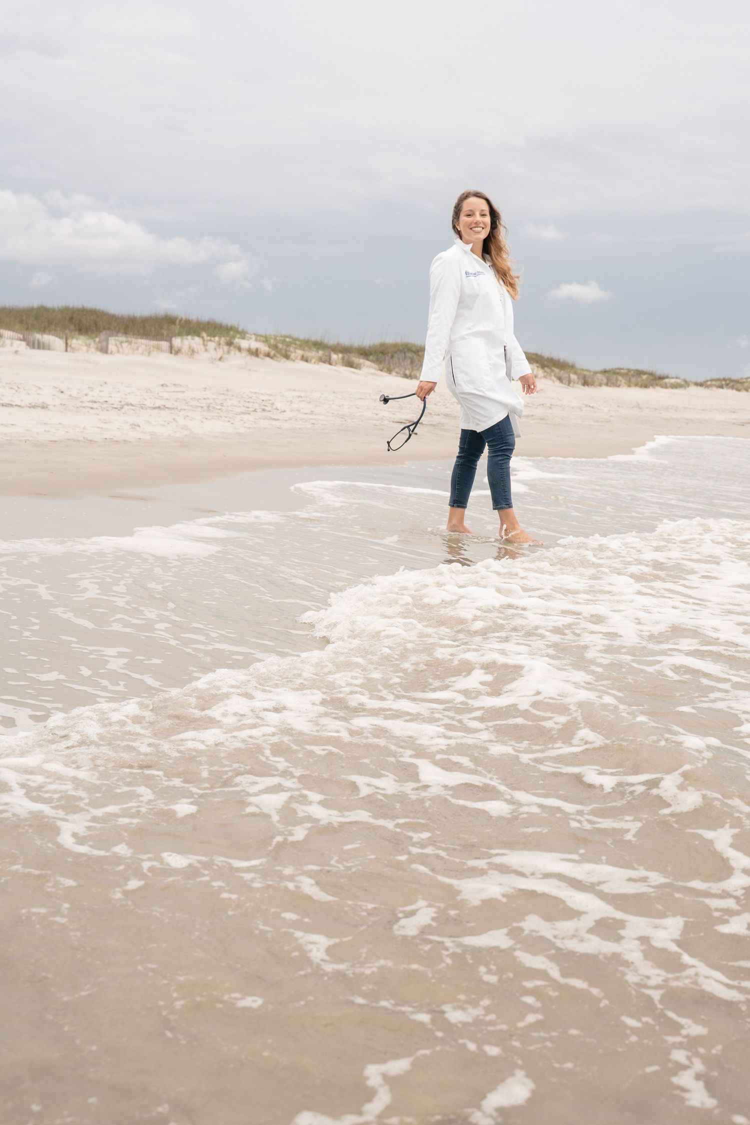 Woman in white shirt and jeans walking barefoot on a sandy beach shore, smiling while holding sunglasses.
