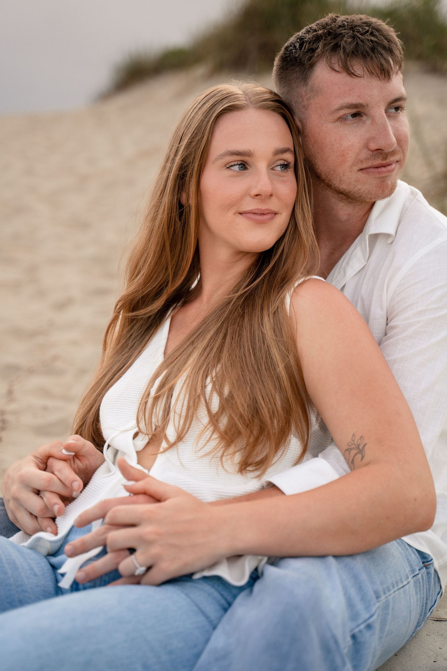 A young couple sits together on a sandy beach, the woman in a white top and jeans leaning back into her partner's embrace.