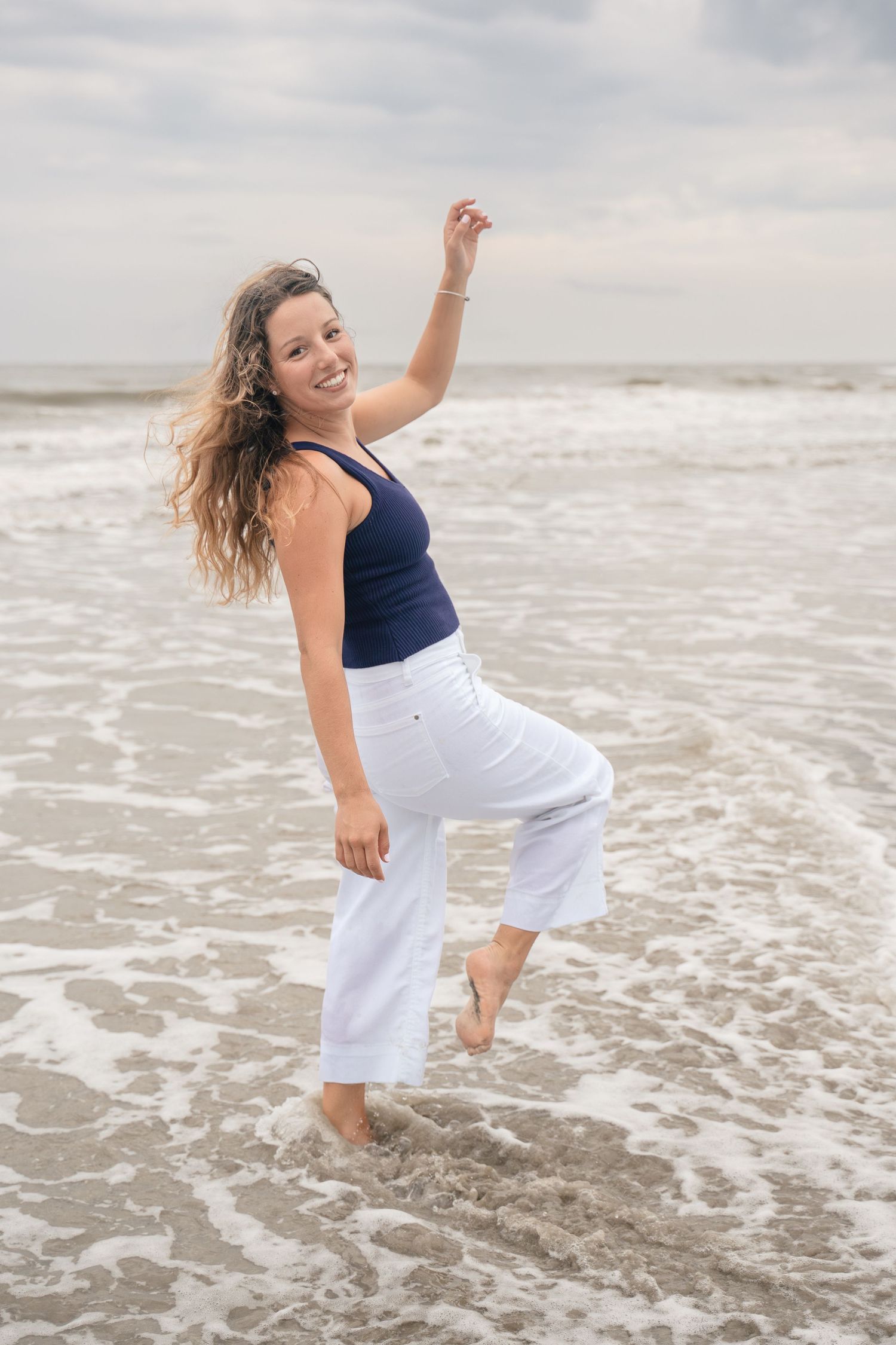 A smiling young woman in white flared pants and navy top dancing barefoot in ocean waves on a beach.