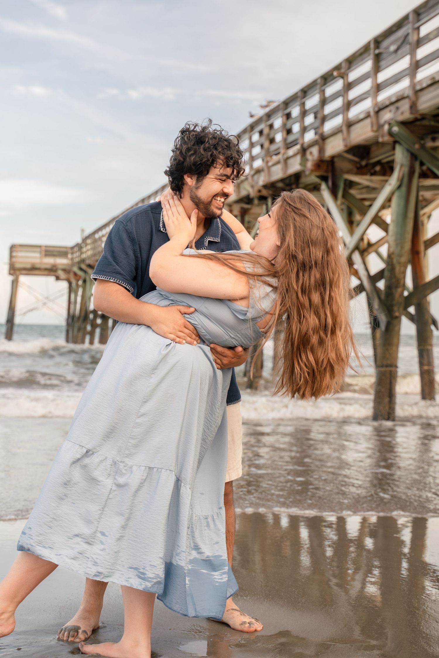 A couple embraces and laughs together on a beach near a wooden pier, with ocean waves in the background.