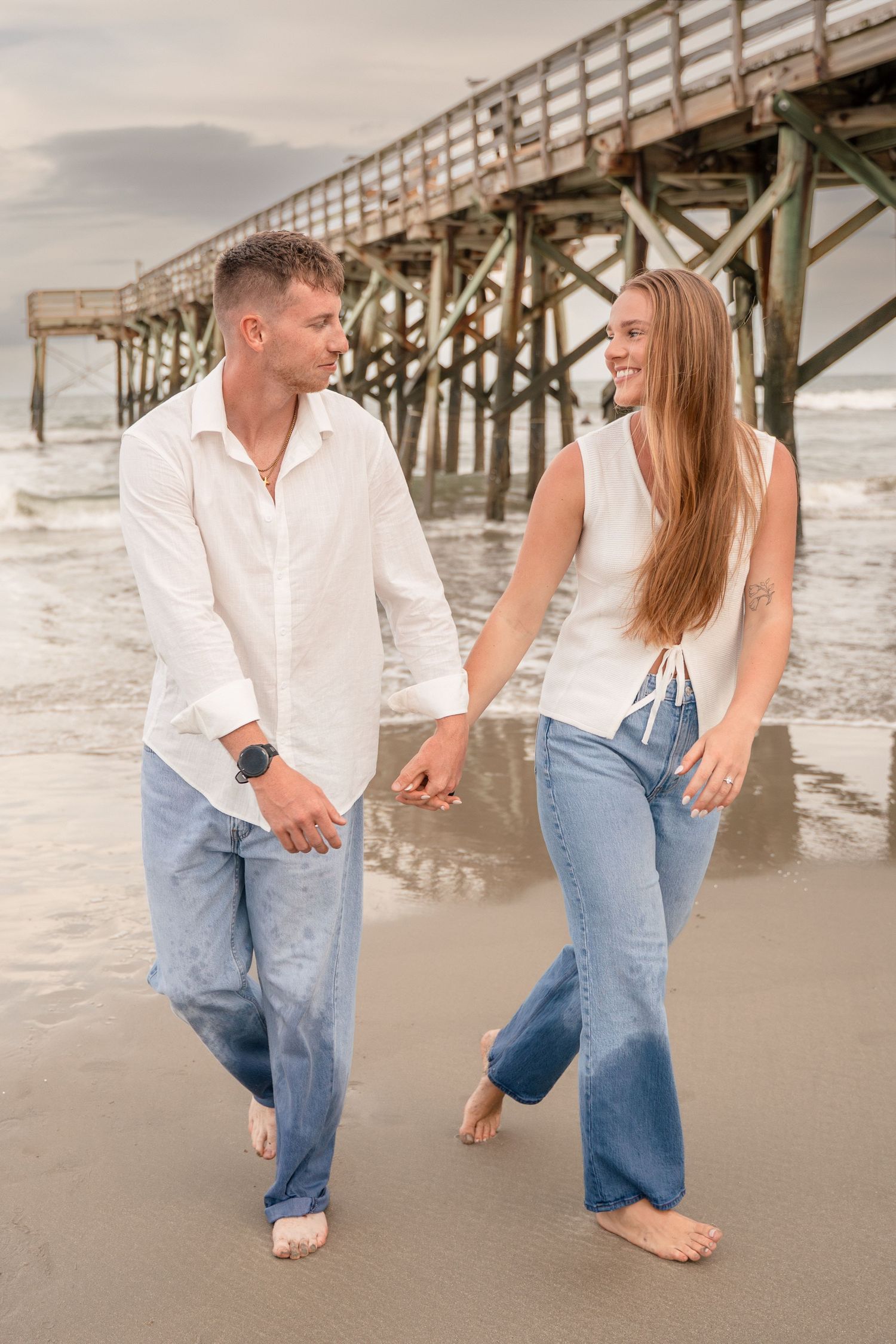 Couple in white tops and jeans holding hands and smiling at each other on a sandy beach by a pier.