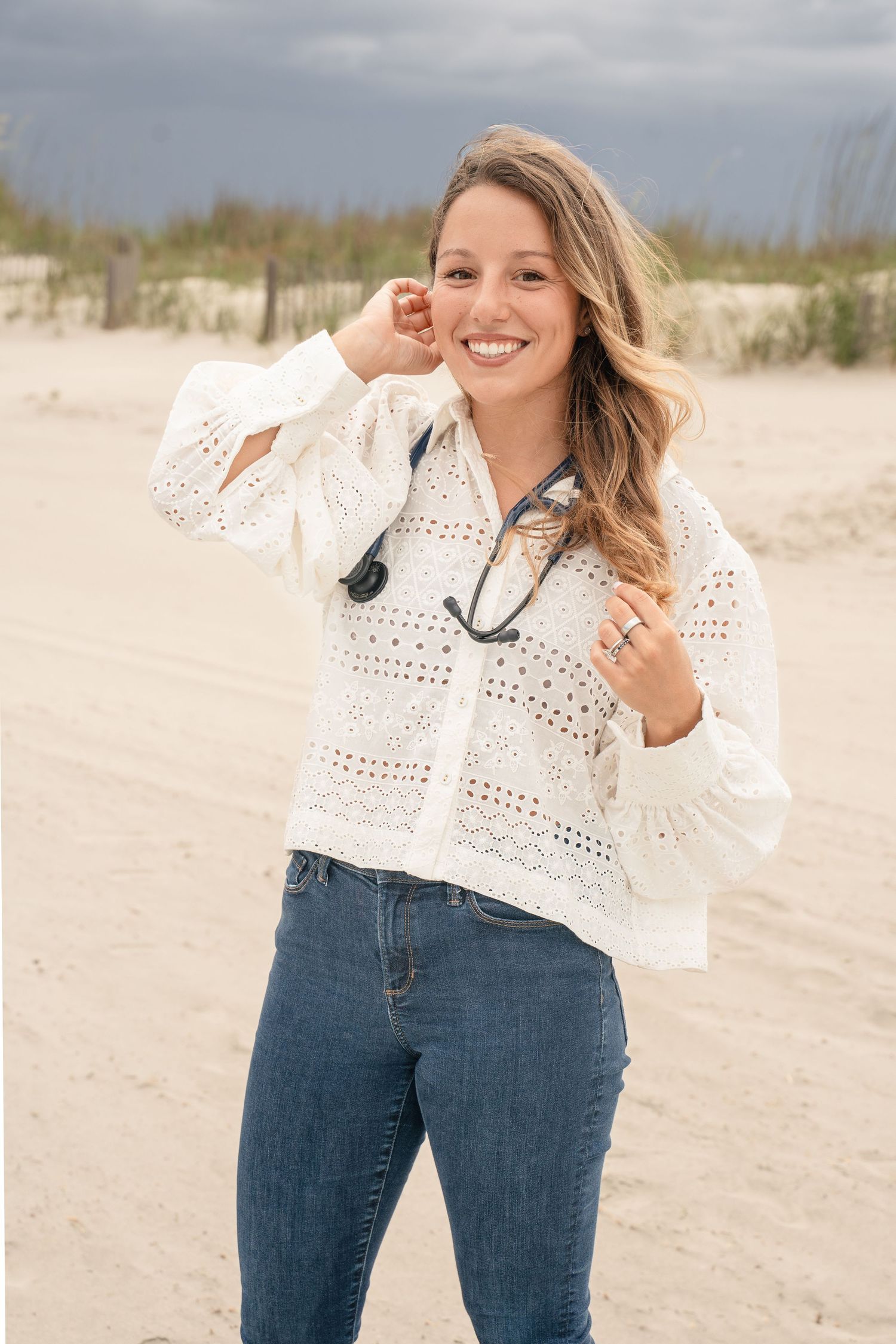 Smiling woman in white lace blouse and jeans holding sunglasses on a sandy beach with stormy skies.