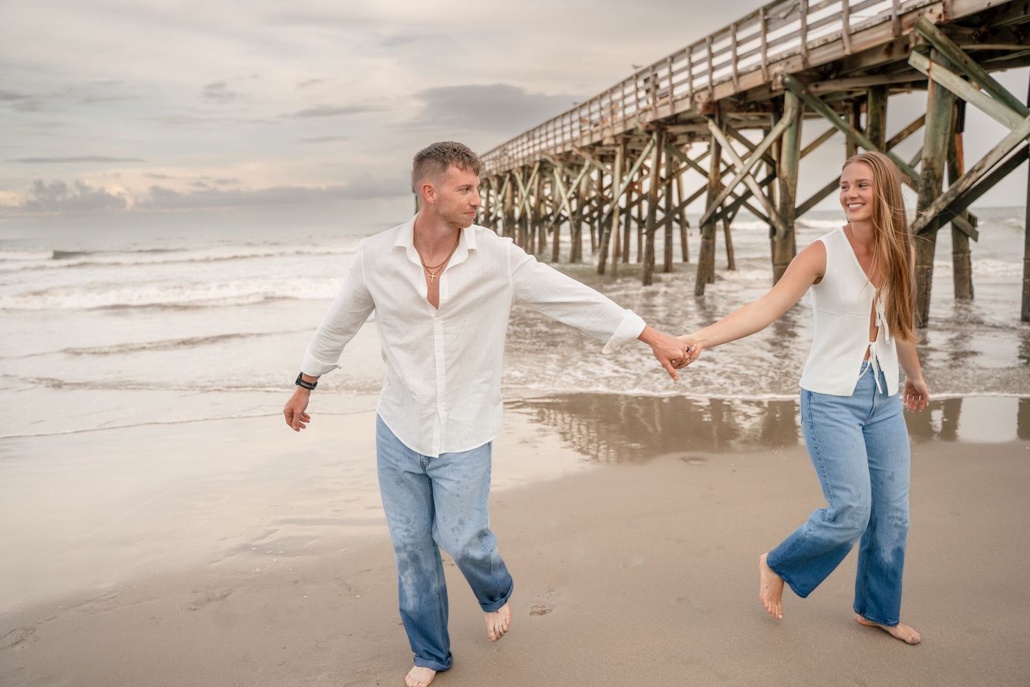 A joyful couple in white shirts and jeans runs hand in hand along a beach near a wooden pier.