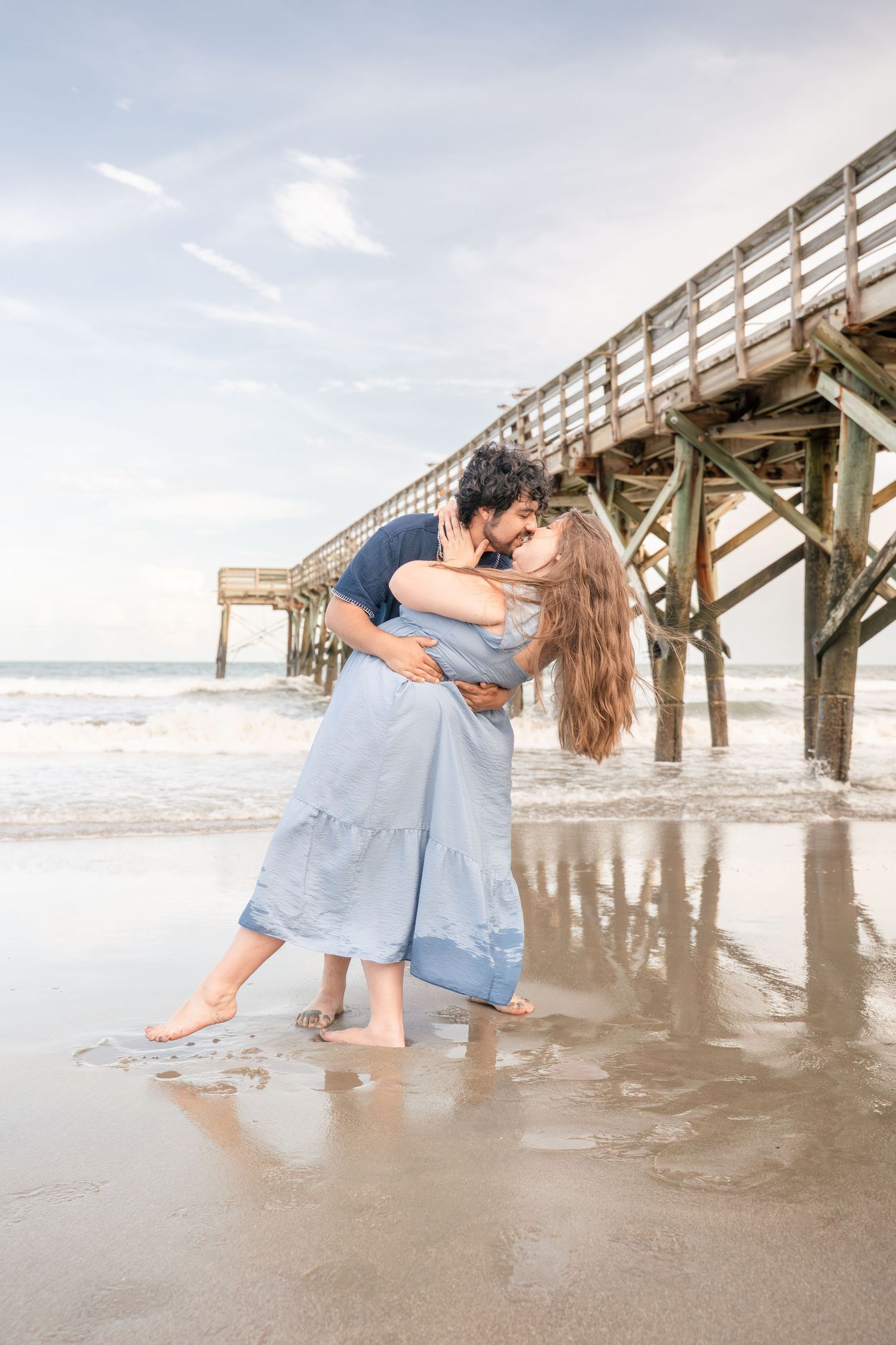 Couple embracing and kissing on a wet sandy beach near a wooden pier with ocean waves in the background.