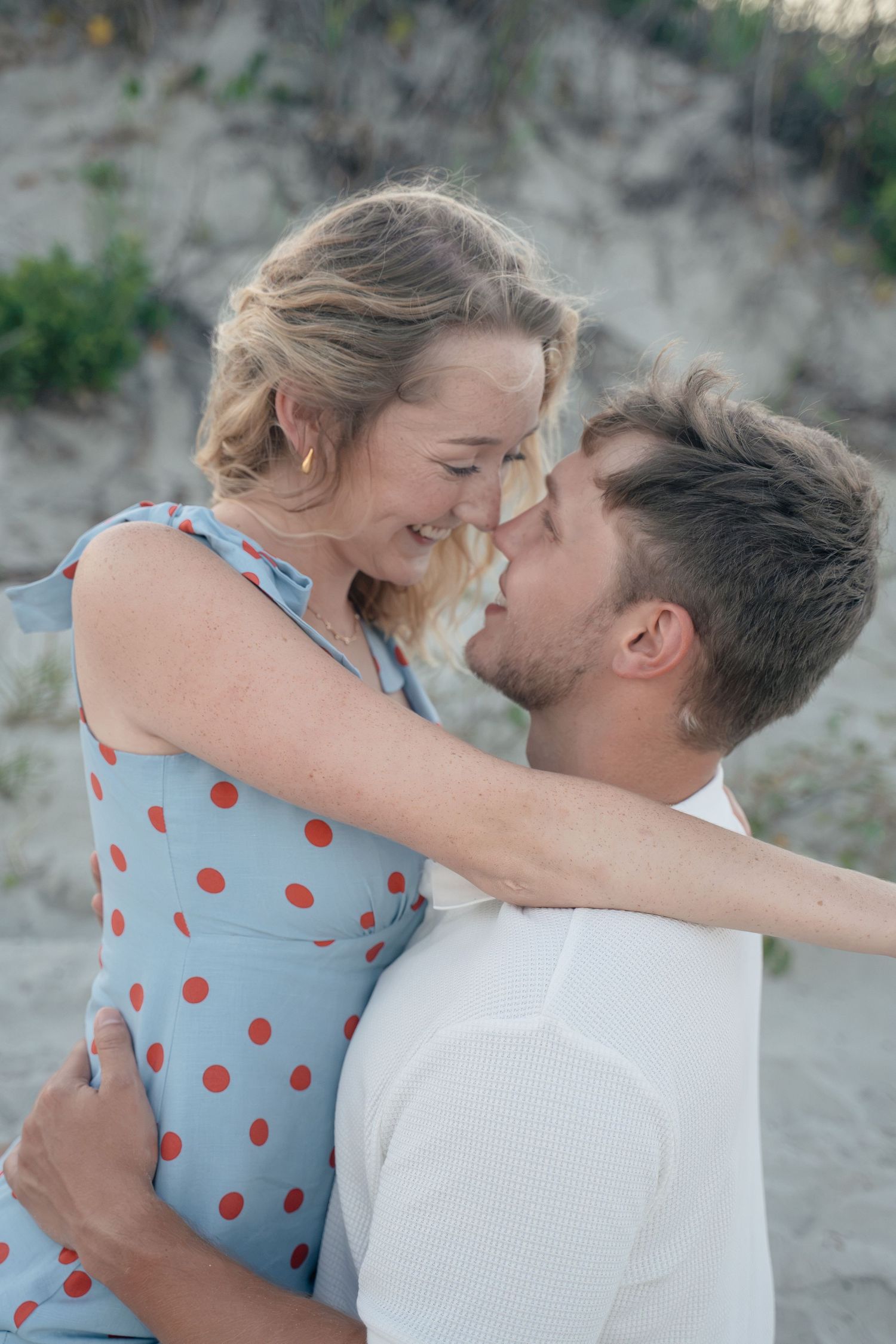 A couple sharing a romantic moment on a sandy beach, the woman in a blue polka dot dress smiling at her partner.