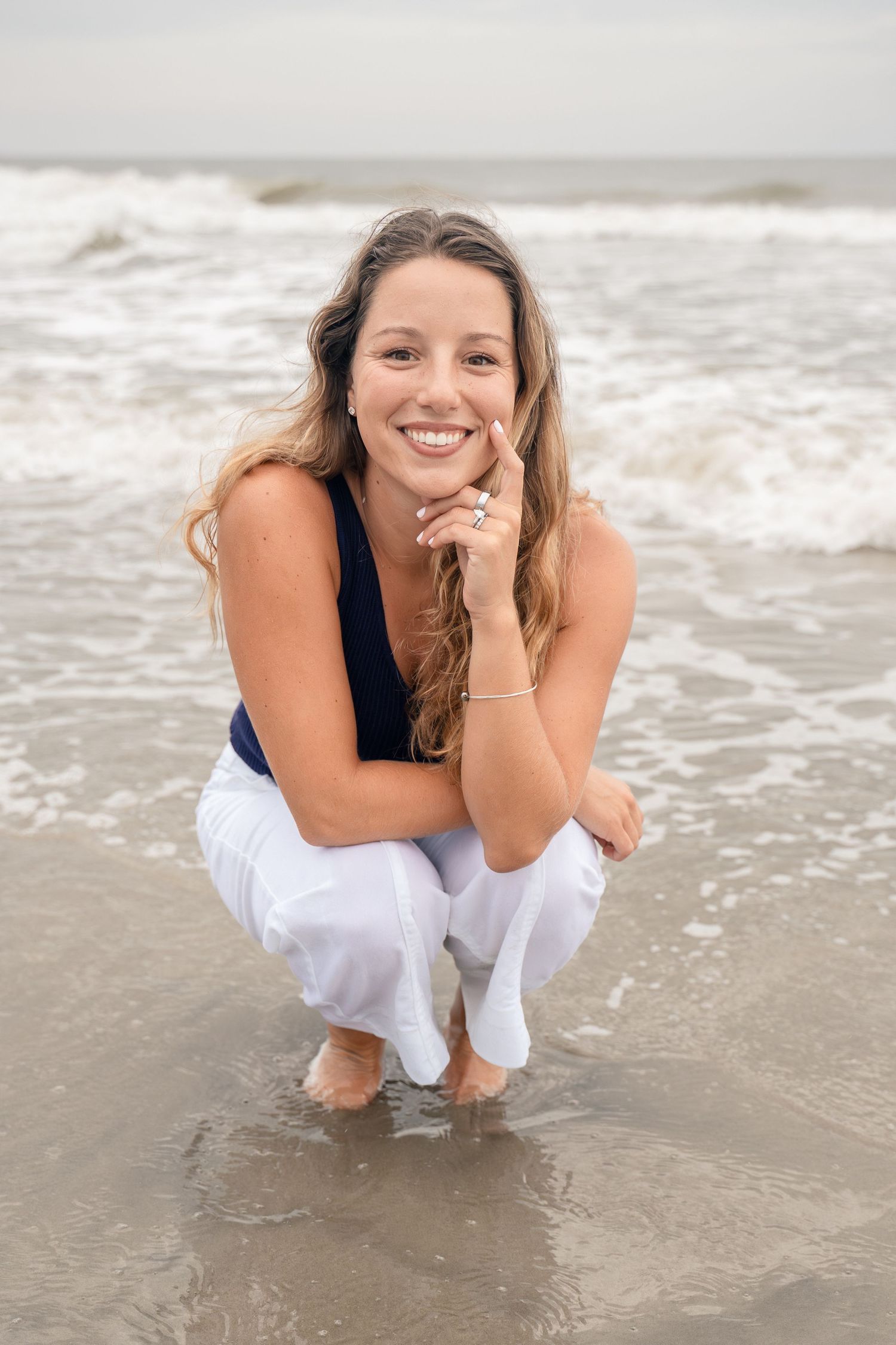 Smiling young woman crouching on a sandy beach in white pants and navy top, with ocean waves in the background.