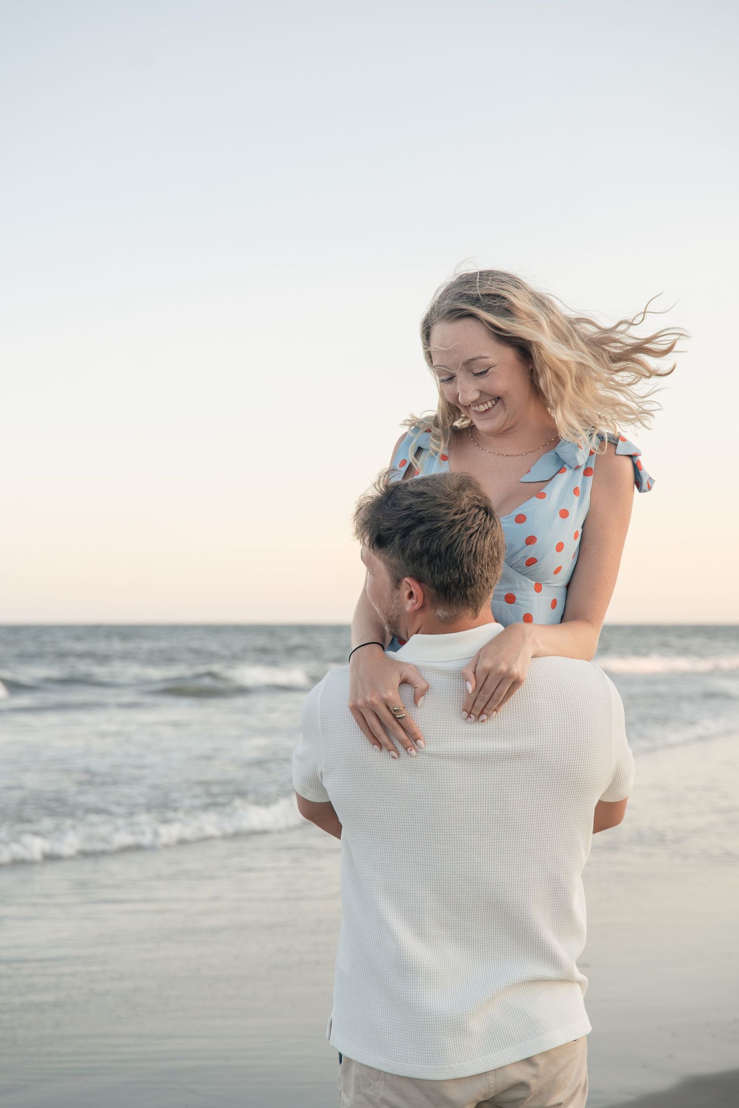 A happy couple embraces on a beach at sunset, woman in polka dot dress lifted by man in white shirt.