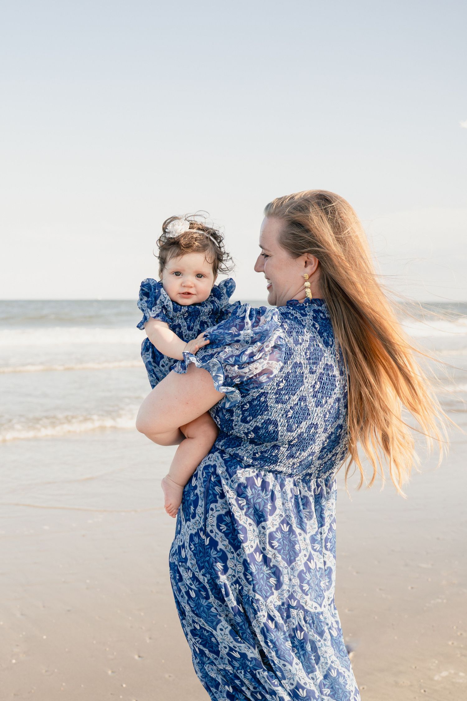 A mother in a blue floral dress holds her baby girl on a sunny beach, both wearing matching outfits.