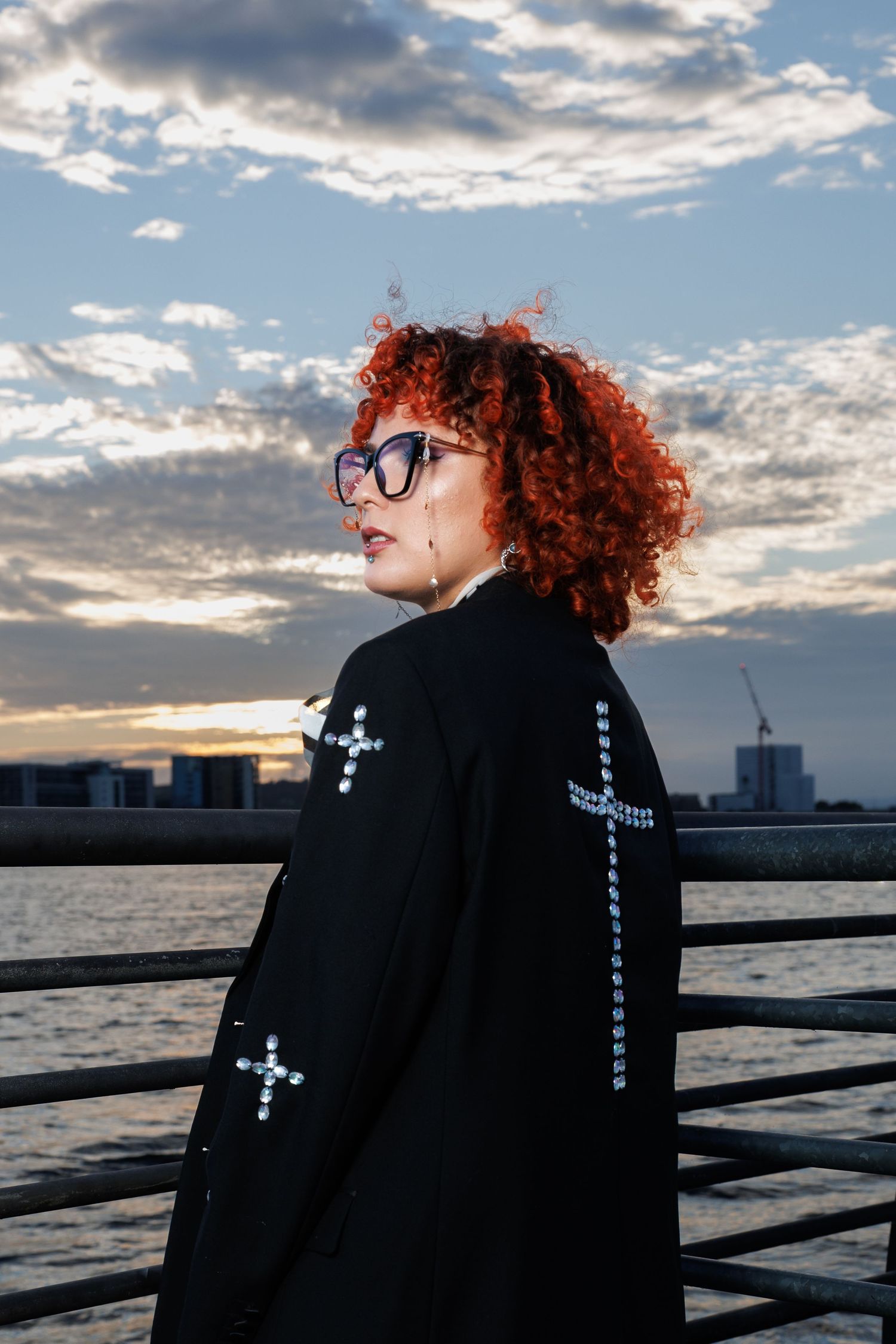 Woman with red curly hair and glasses wearing a black cross-embellished jacket, standing by waterfront at sunset.