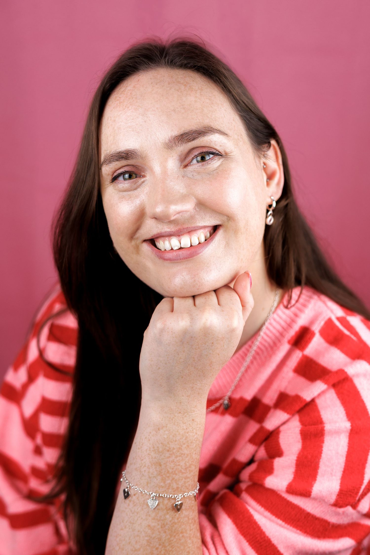 Smiling brunette woman with freckles poses against a pink background wearing a striped top.