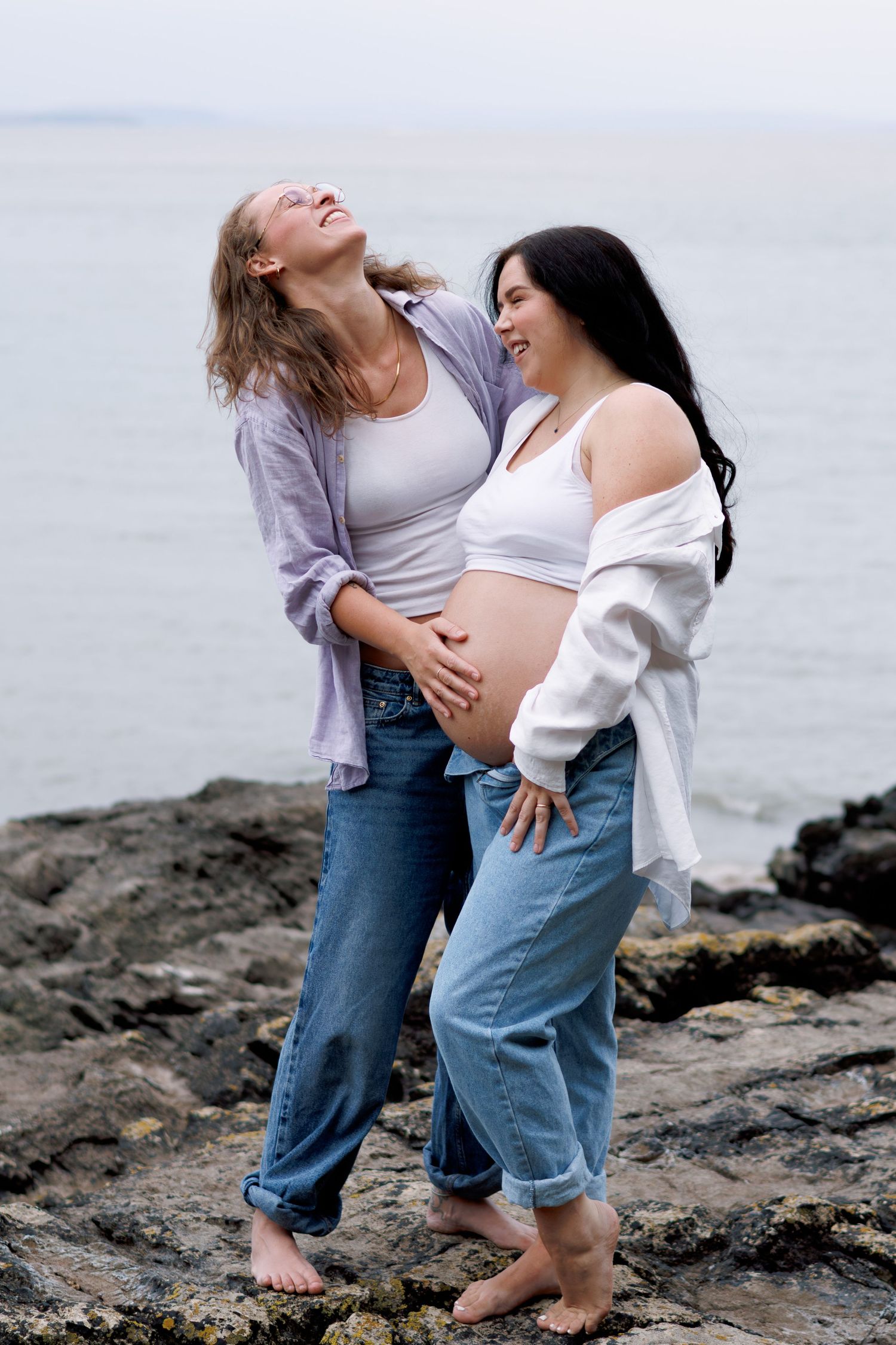Two women laughing together on rocky beach, one pregnant, both wearing jeans and open shirts by the ocean.