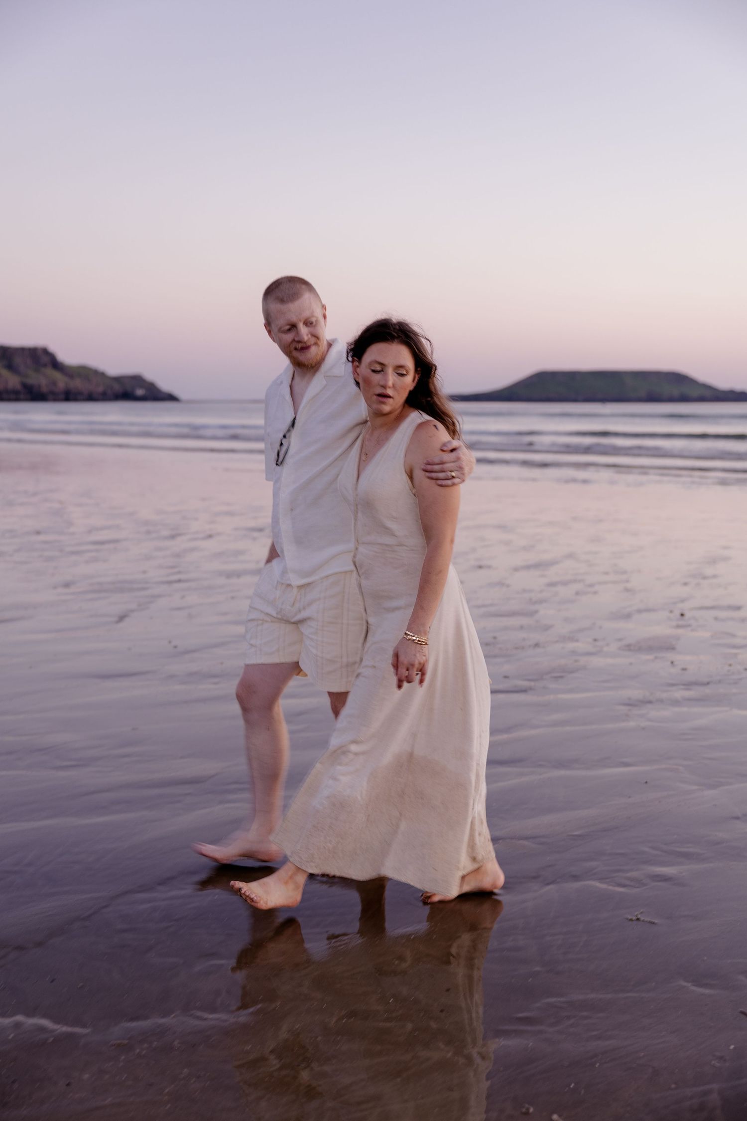 Couple walking barefoot on a beach at dusk, woman in flowing white dress, man in white shirt and shorts.