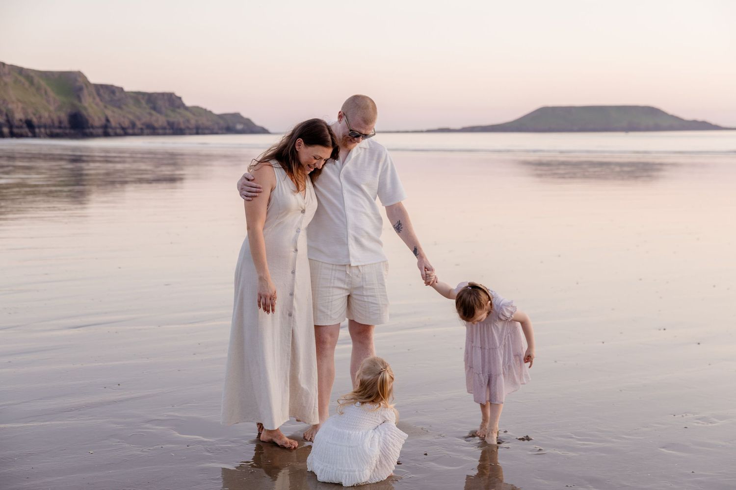 Family of four enjoying a serene beach walk at sunset, with two young girls and parents dressed in white.