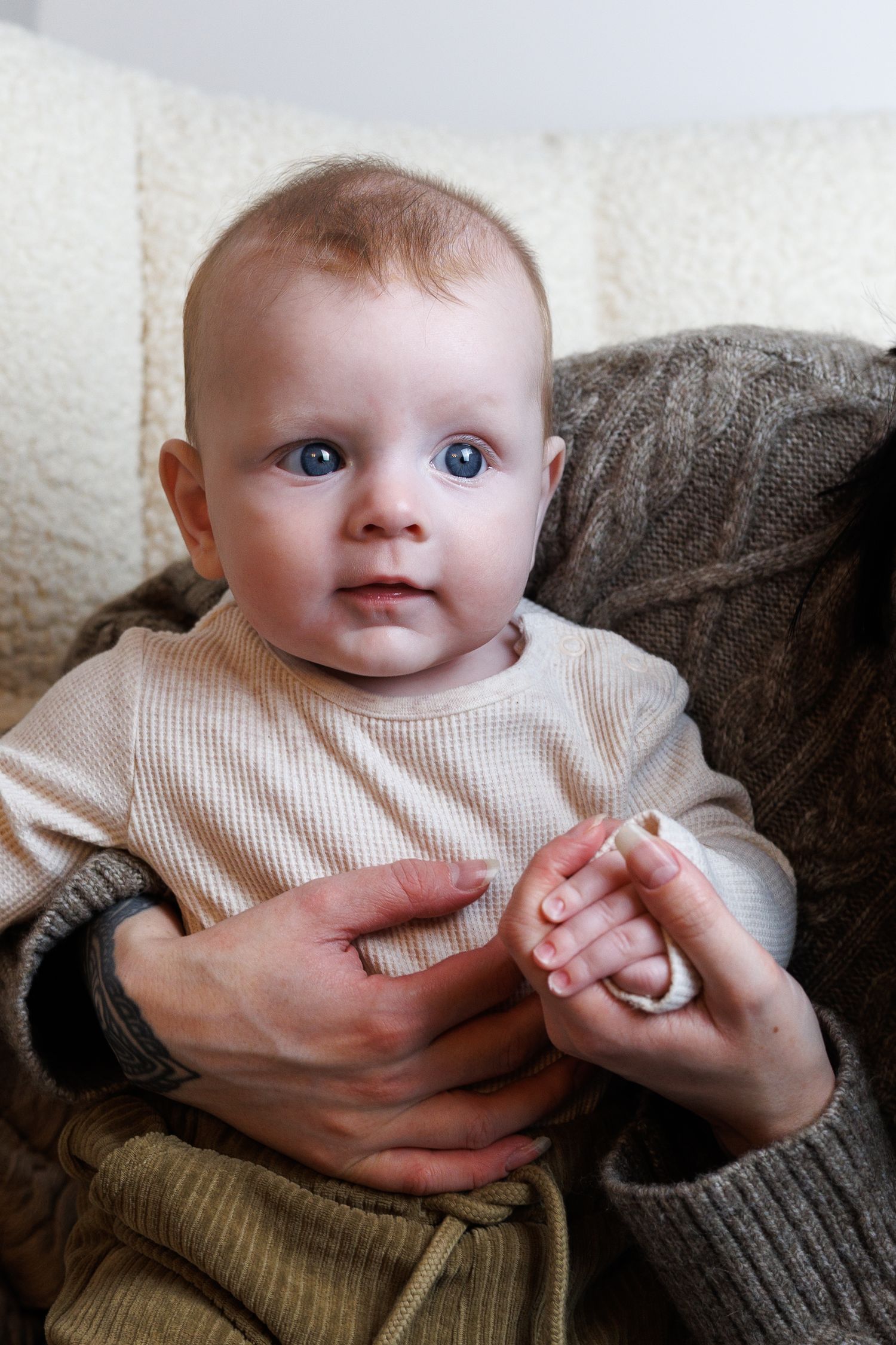 Adorable blue-eyed baby in beige outfit sits supported by adult hands on a cozy chair.