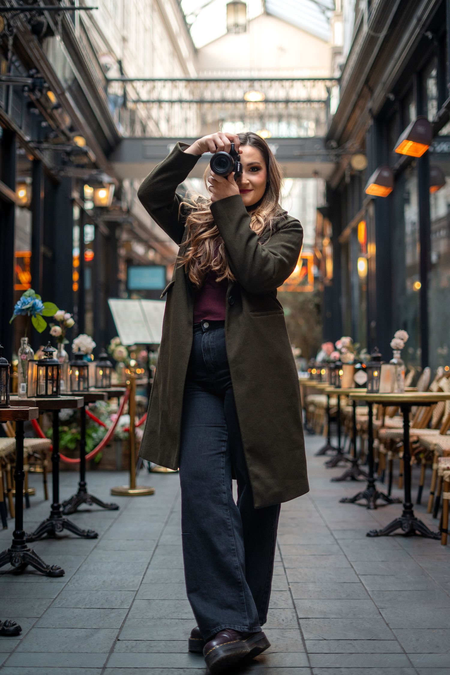 Woman in olive coat photographing with DSLR camera in elegant indoor arcade cafe.