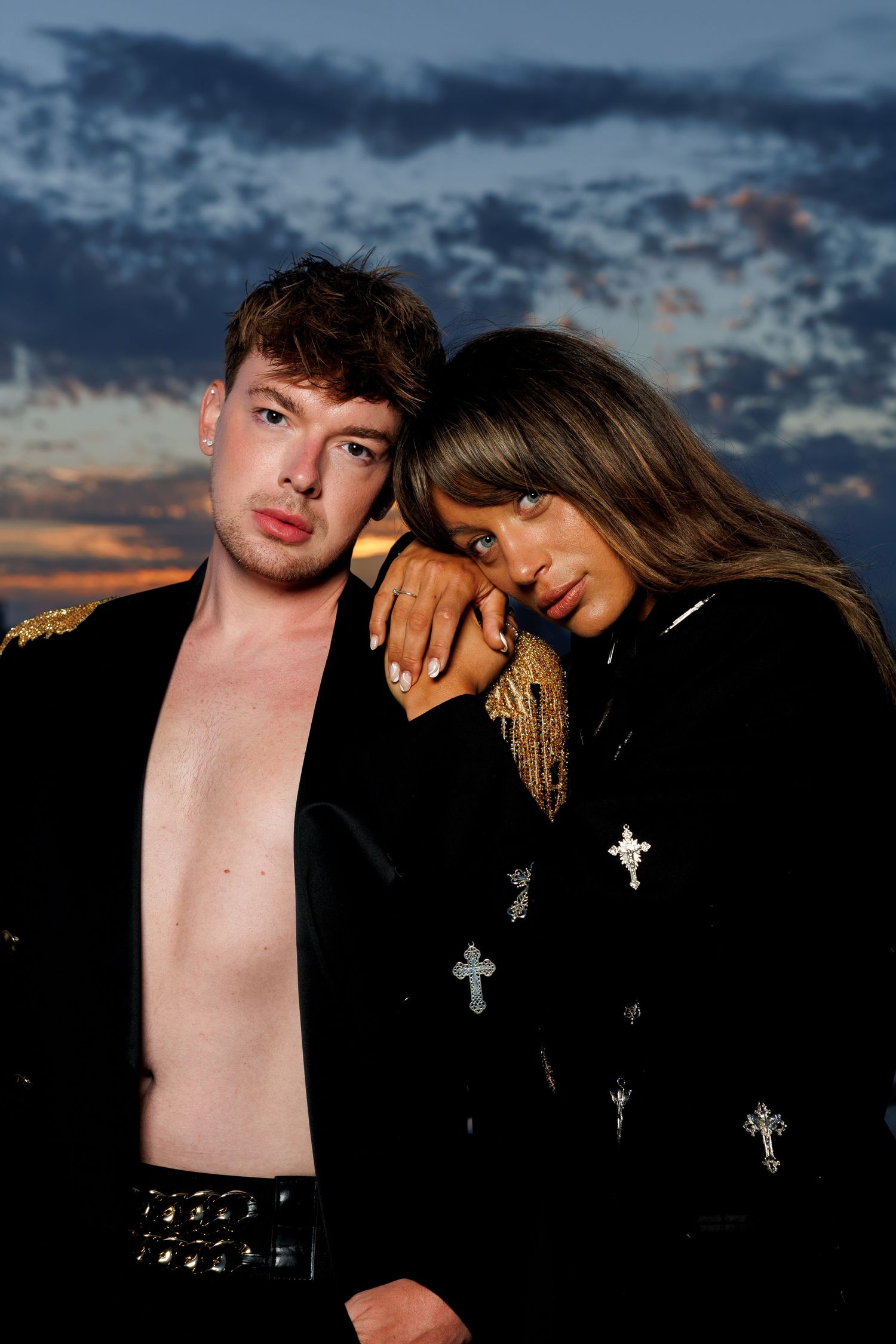 Two people in black velvet jackets with cross embroidery pose dramatically against a sunset sky.