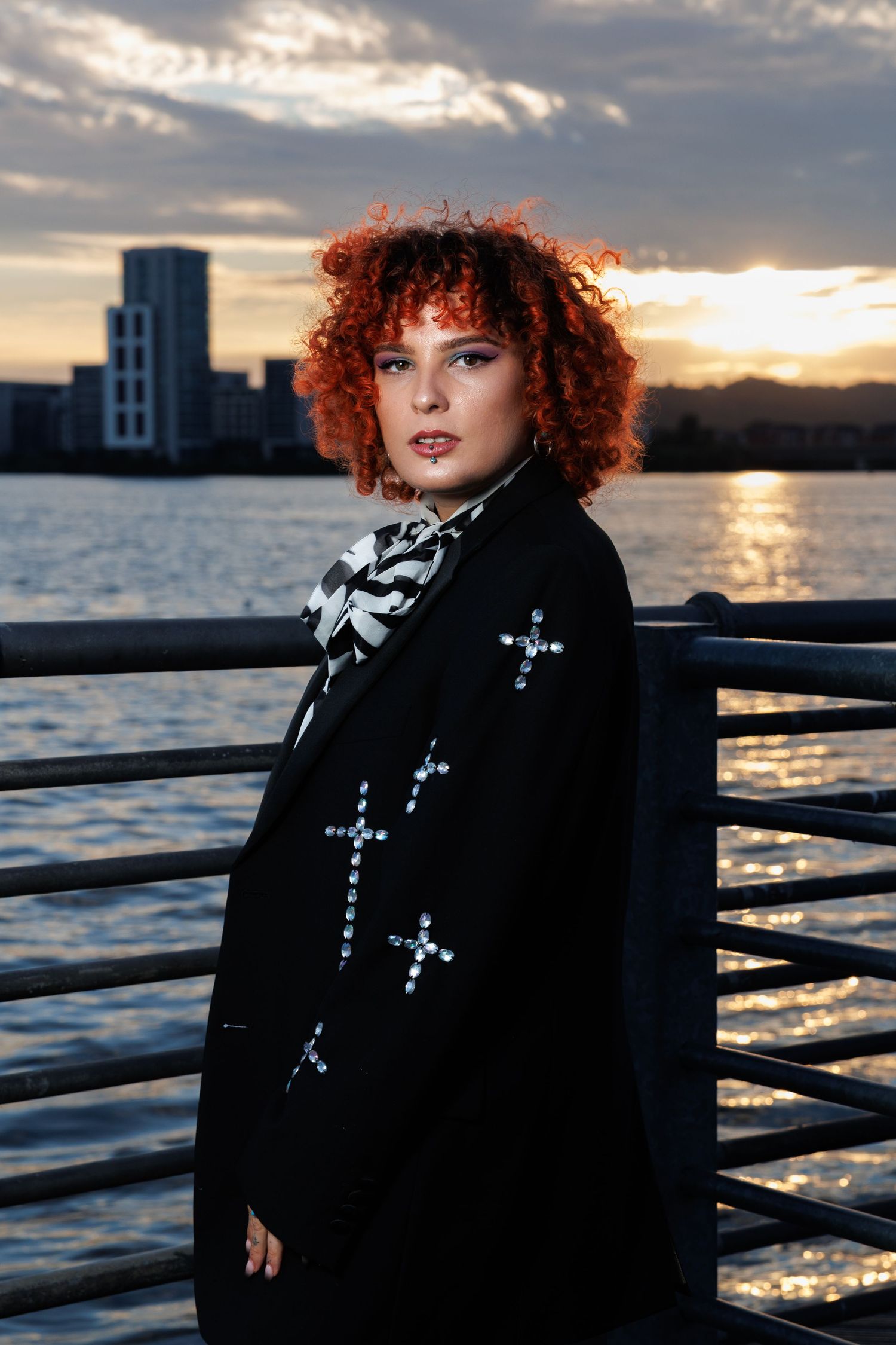 Young woman with curly red hair wearing a black cross-embellished blazer poses by waterfront railing at sunset.