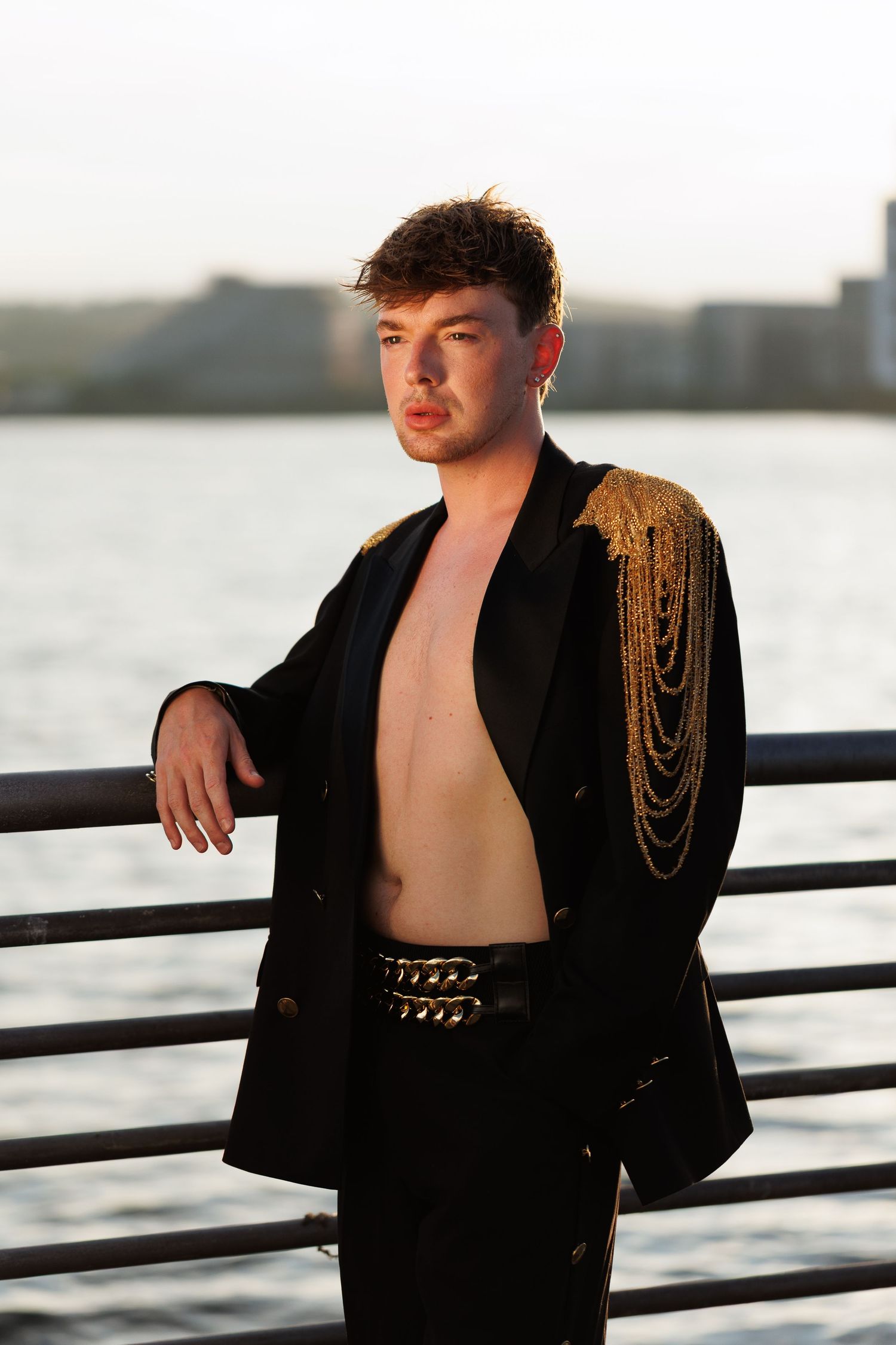 Young man in open black blazer with gold chain epaulettes poses by waterfront railing at sunset.