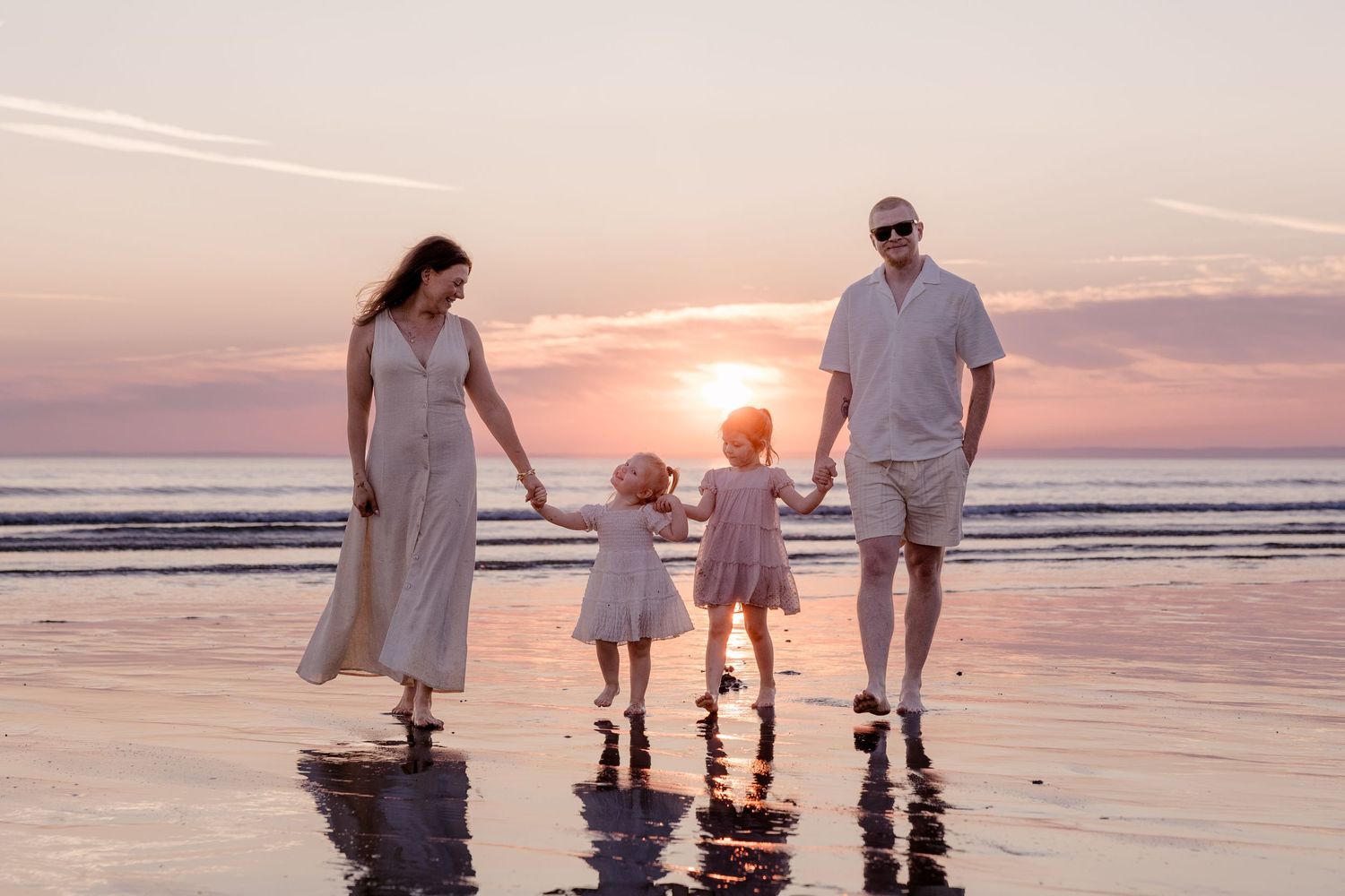 Family of four holding hands walking on a beach at sunset in white outfits.