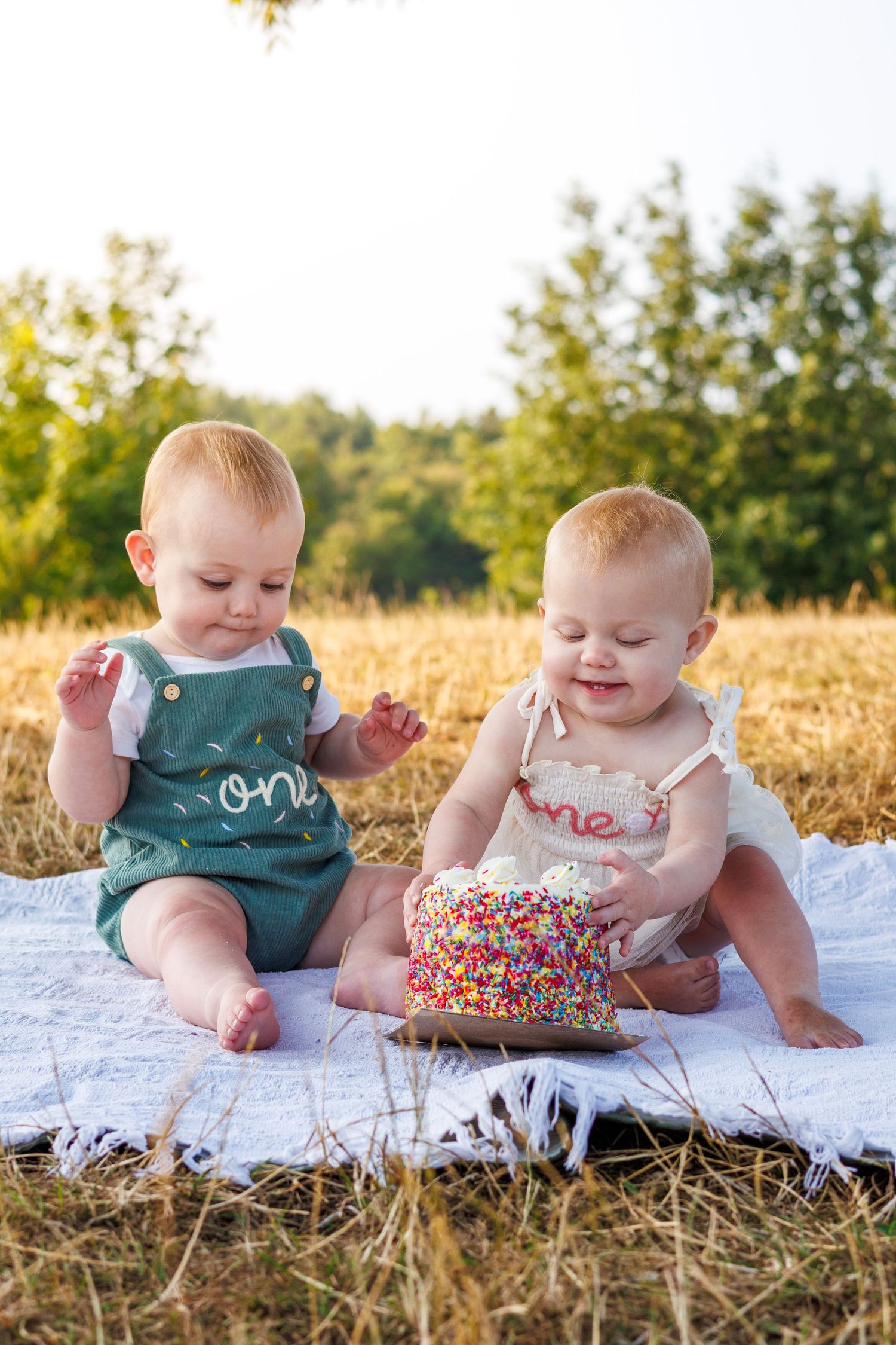 Two babies in matching outfits celebrate their first birthday with a colorful sprinkle cake outdoors.